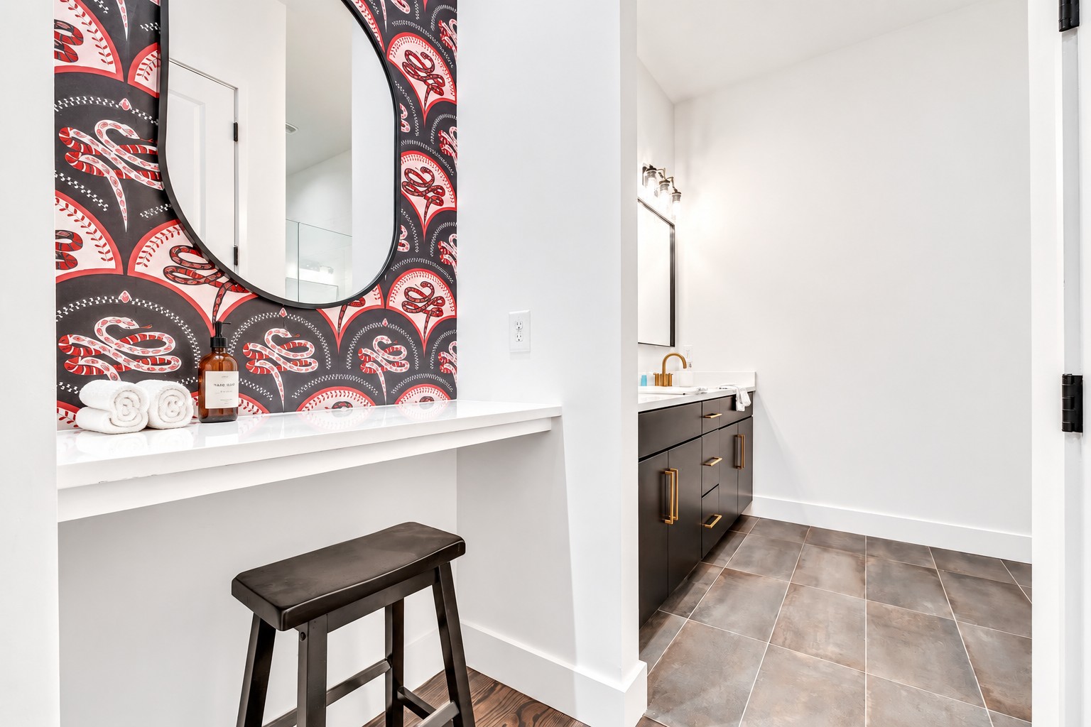 Bathroom vanity with bold patterned wallpaper, an oval mirror, rolled towels, and dark cabinets with brass hardware.