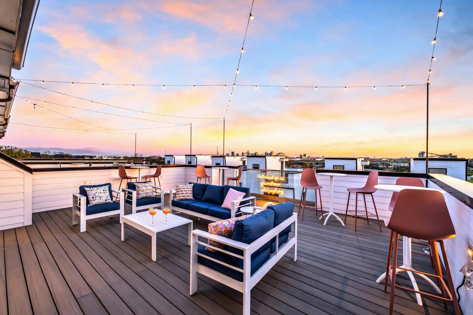 Furnished rooftop deck with string lights, lounge seating, and a distant city skyline at sunset.
