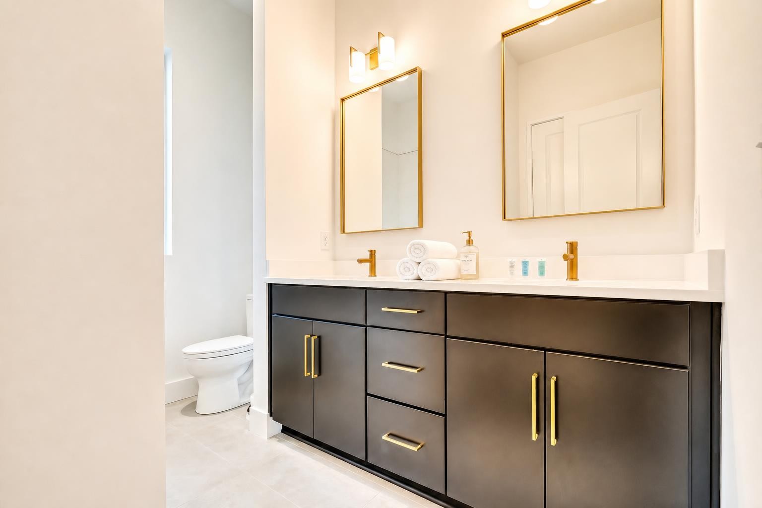 Bathroom with a dark double vanity, gold fixtures, two framed mirrors, and a toilet.