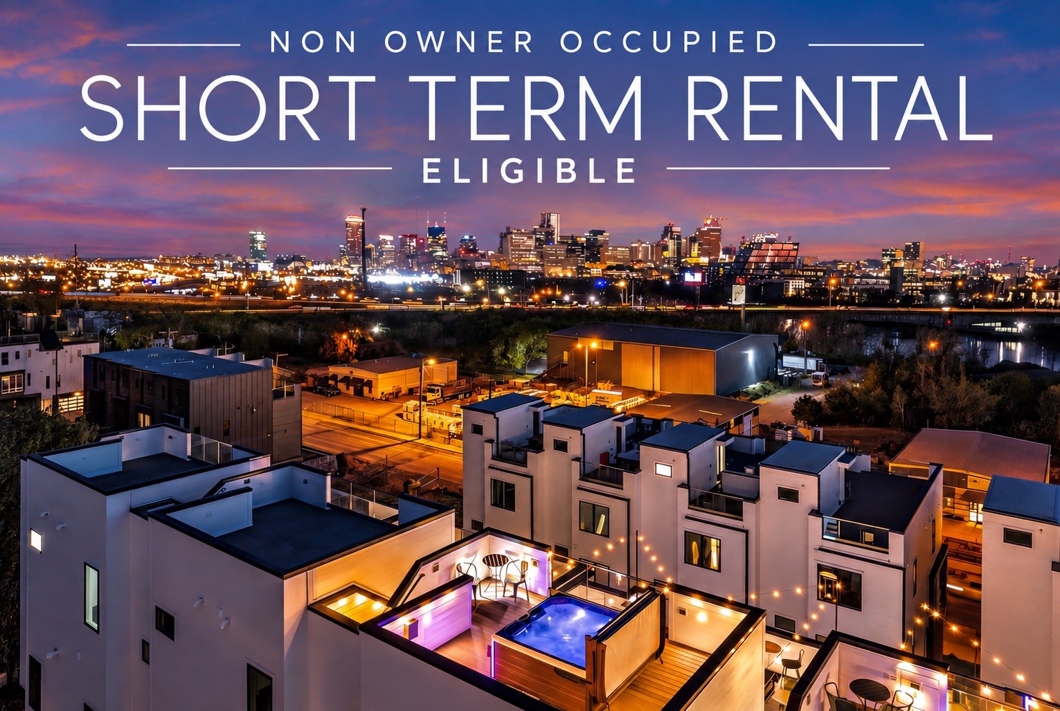 Modern townhomes with rooftop decks and a pool overlook the illuminated downtown skyline at dusk.