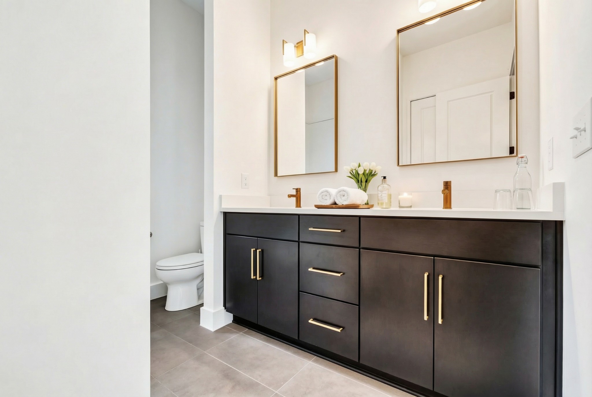 Bathroom with a dark double vanity, brass fixtures, two mirrors, and a partly screened toilet.