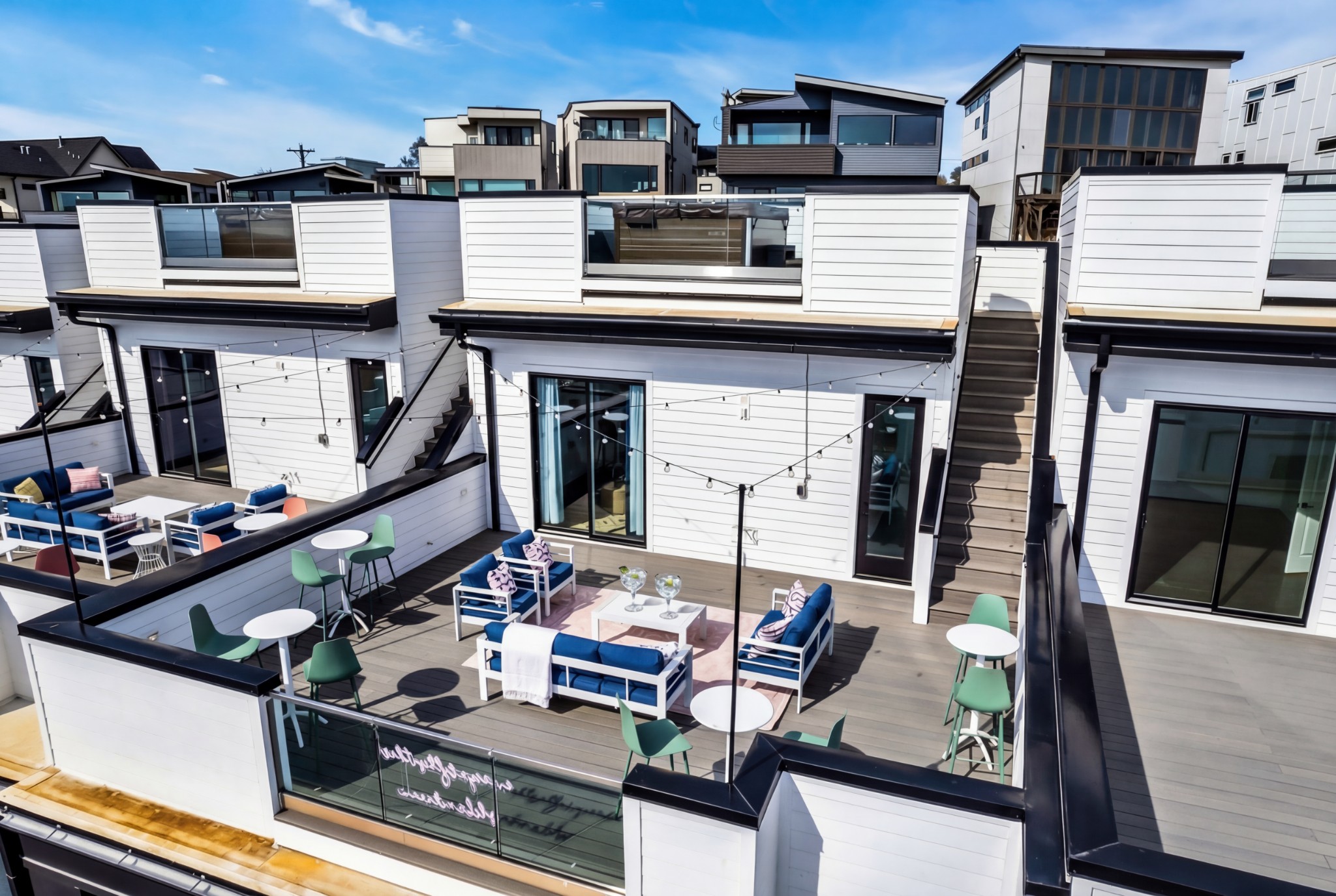 Rooftop deck with blue lounge seating, café tables, string lights, and stairs between modern white buildings.