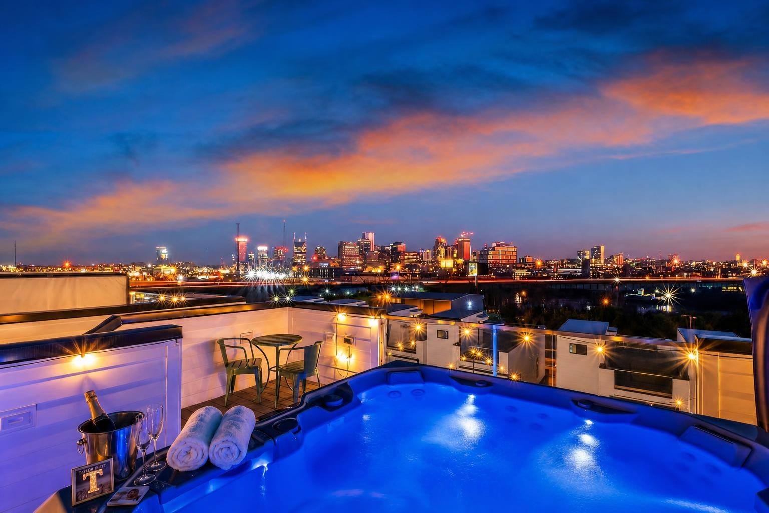 Rooftop hot tub and terrace overlook an illuminated city skyline beneath blue and orange dusk clouds.