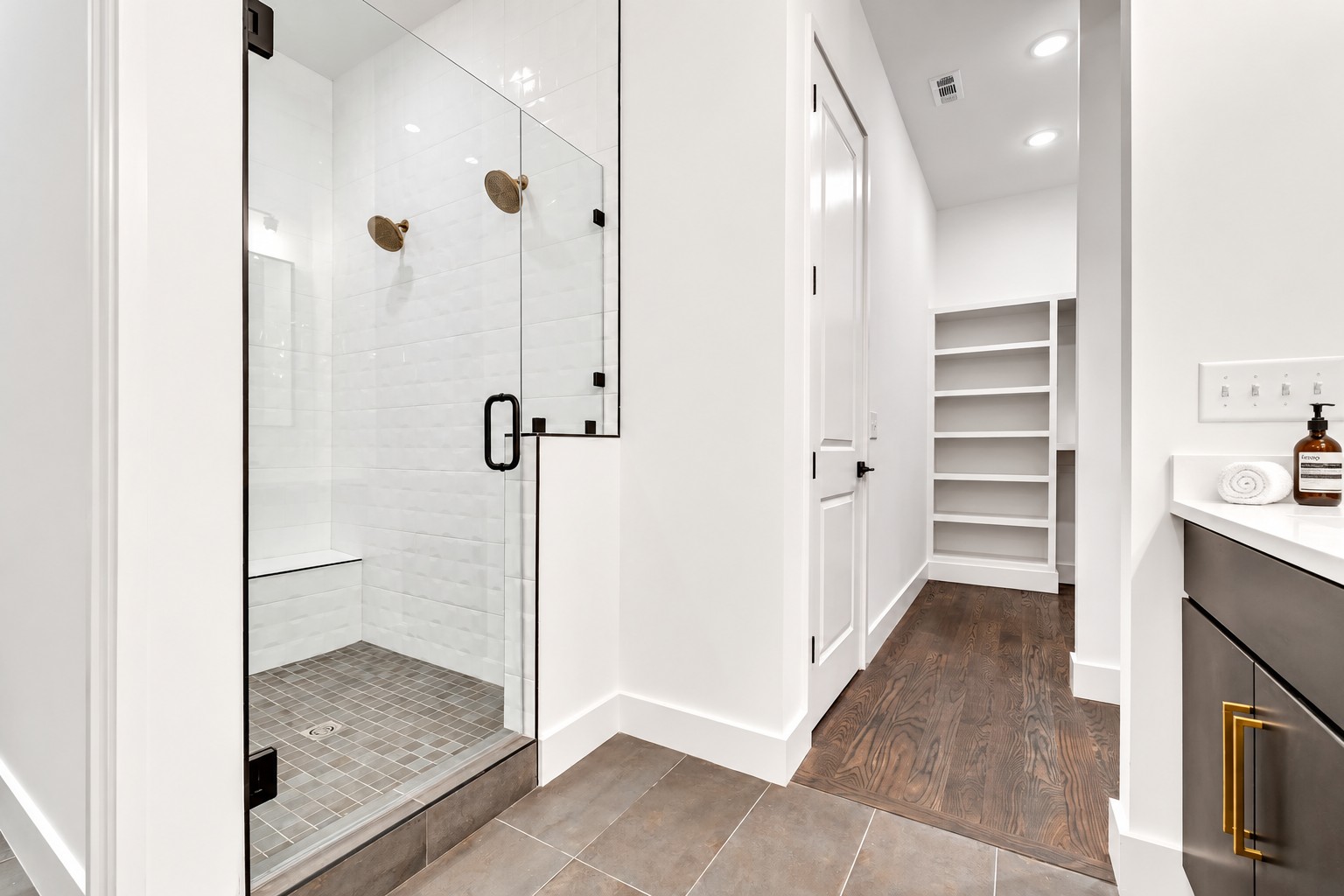 Modern bathroom with a glass walk-in shower, dual brass showerheads, dark vanity, and built-in shelving.