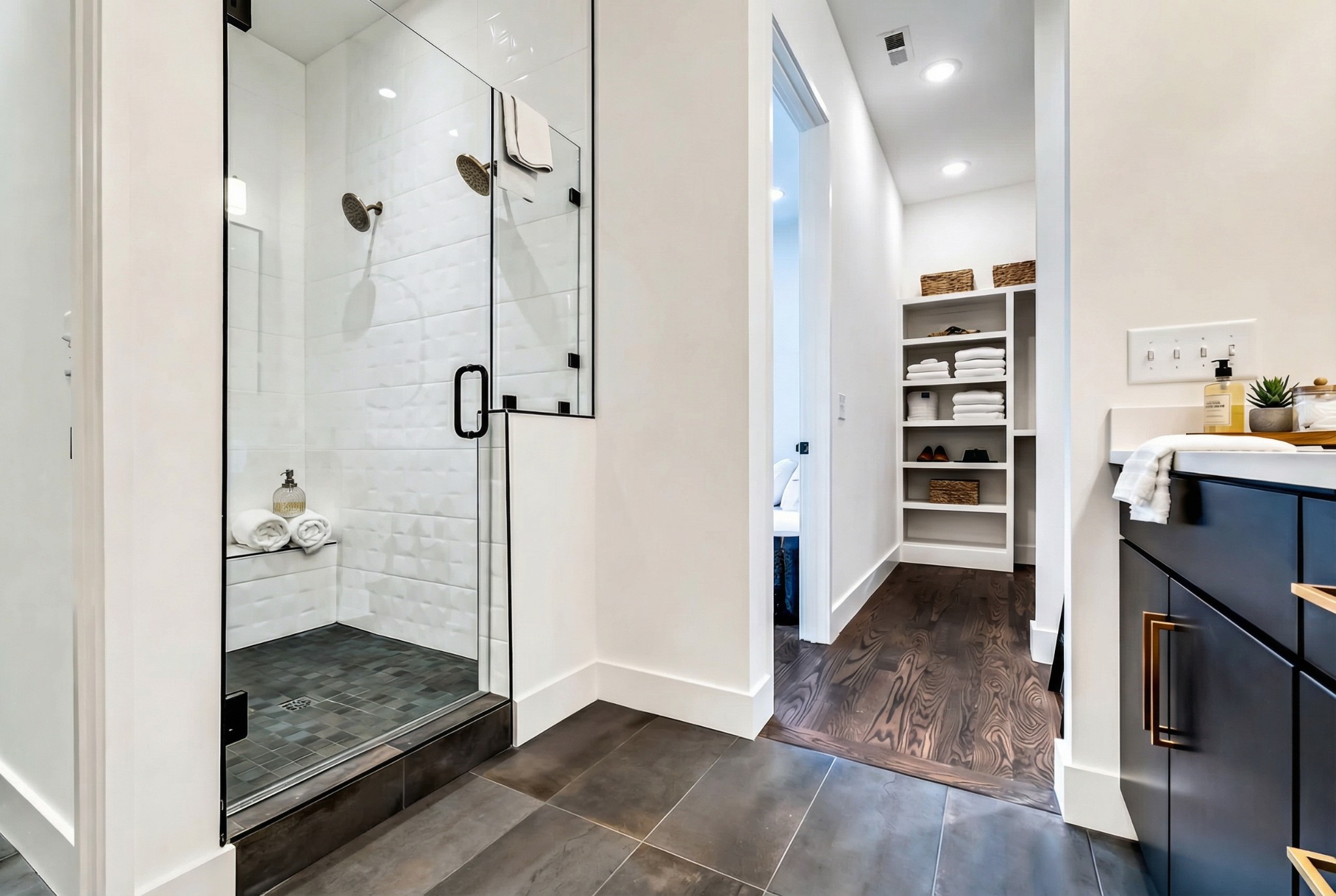 Modern bathroom with glass walk-in shower, navy vanity, and open dressing area with built-in shelves.