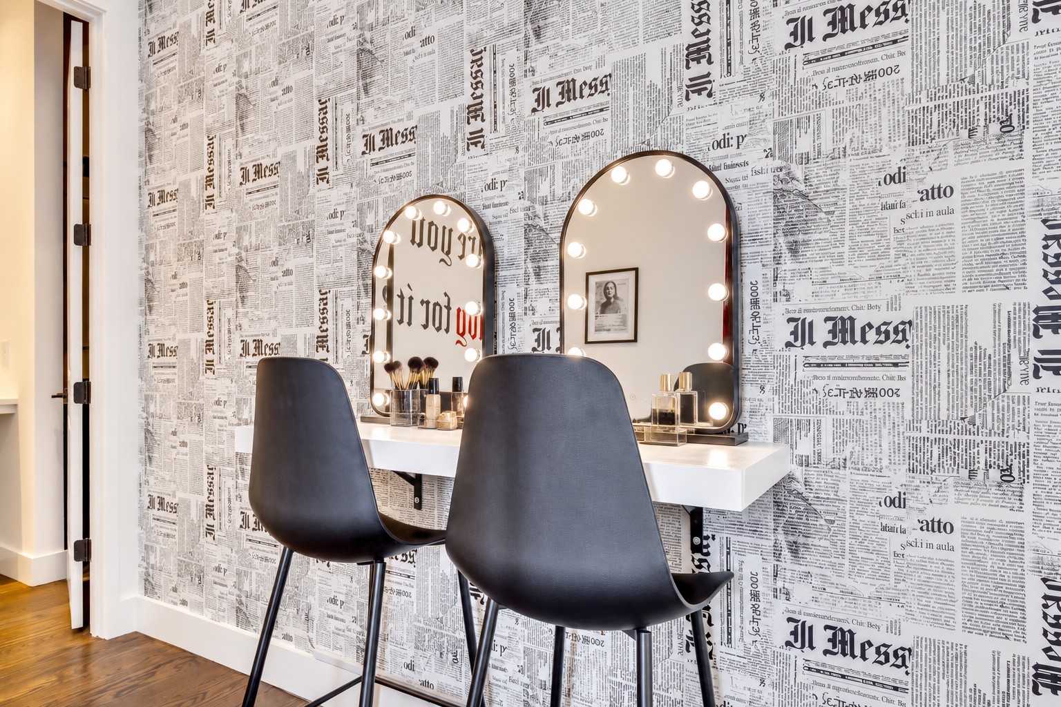 Dressing area with a white vanity, bulb-framed mirrors, black chairs, and newspaper-print wallpaper.