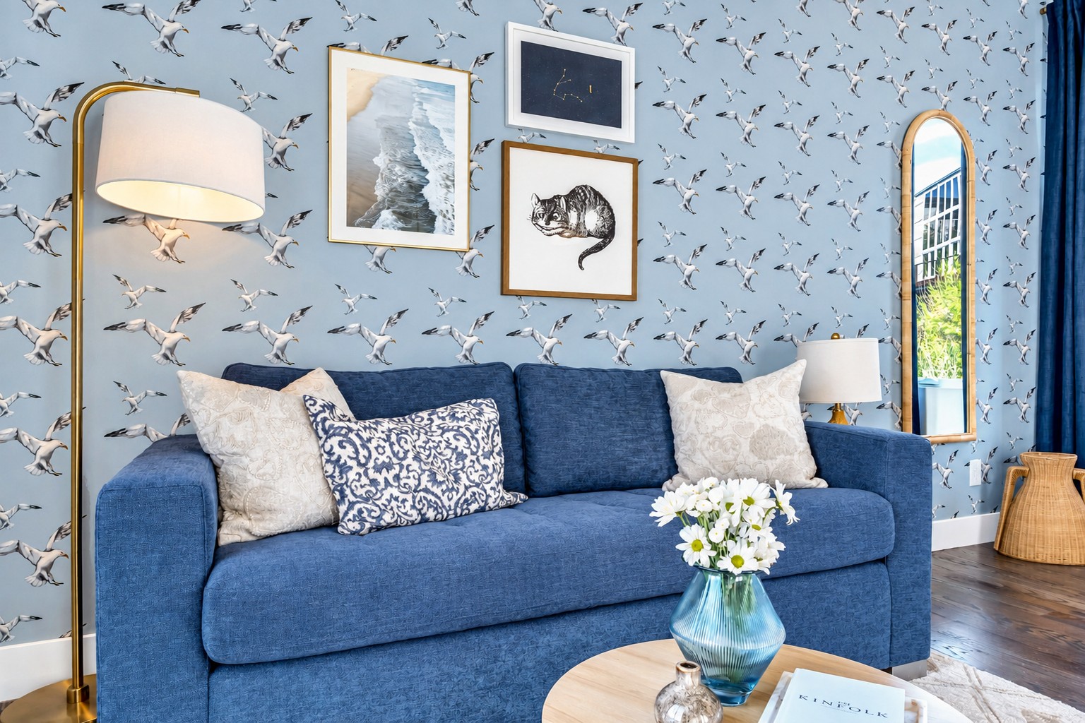 Living room with a blue sofa, bird-print wallpaper, framed art, and a tall arched mirror.