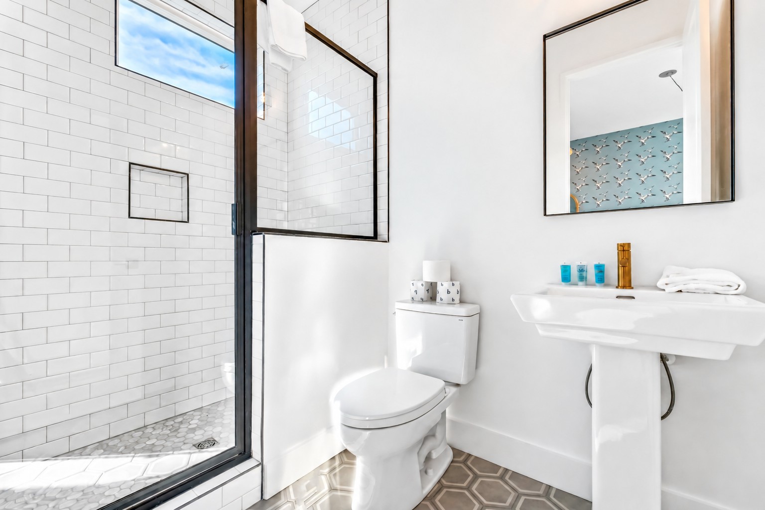 Bright bathroom with a subway-tile shower, pedestal sink, brass faucet, and patterned floor.