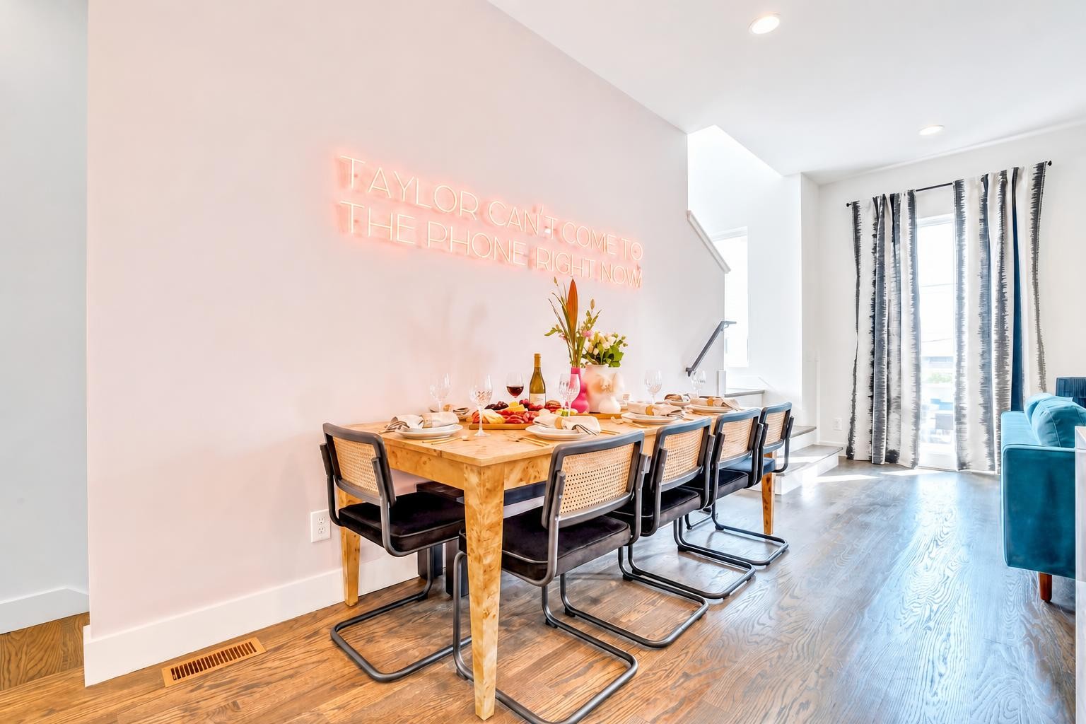 Dining area with a dinner table, cane-back chairs, glowing neon sign, and tall windows.