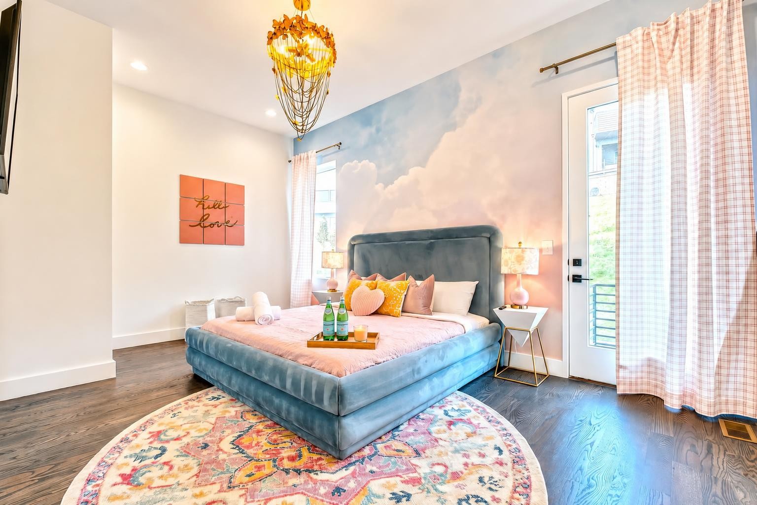 Staged bedroom with a blue upholstered bed, cloud mural, plaid curtains, and colorful round rug.