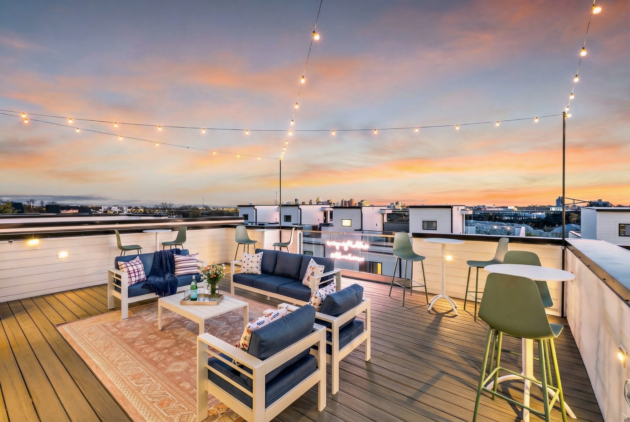 Rooftop deck with lounge seating and string lights overlooking a city skyline at dusk.