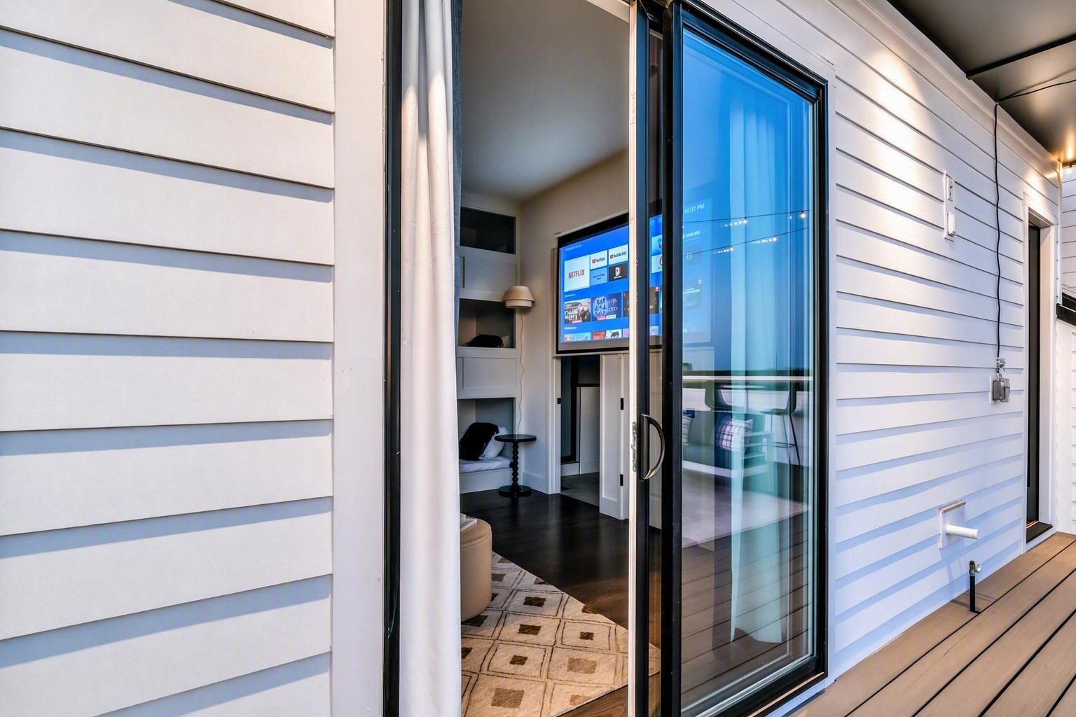 Open sliding glass door from a wooden deck reveals a furnished room with a television and patterned rug.