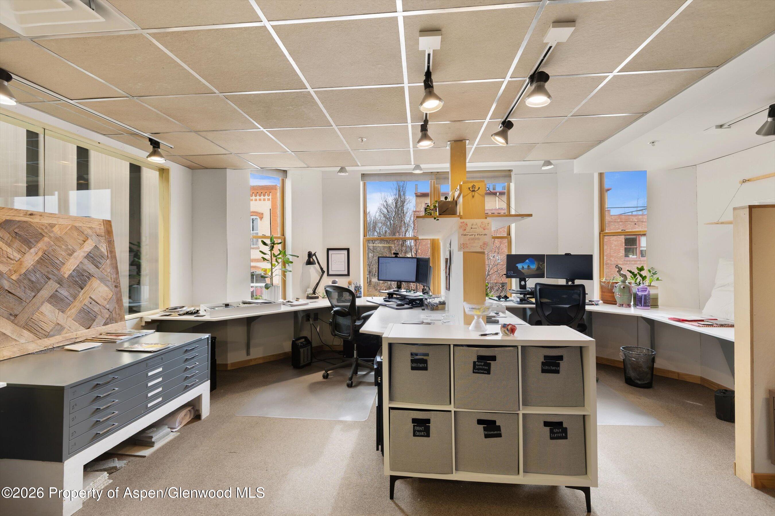 Office workspace with multiple desks, computers, sample drawers, and tall downtown-facing windows.
