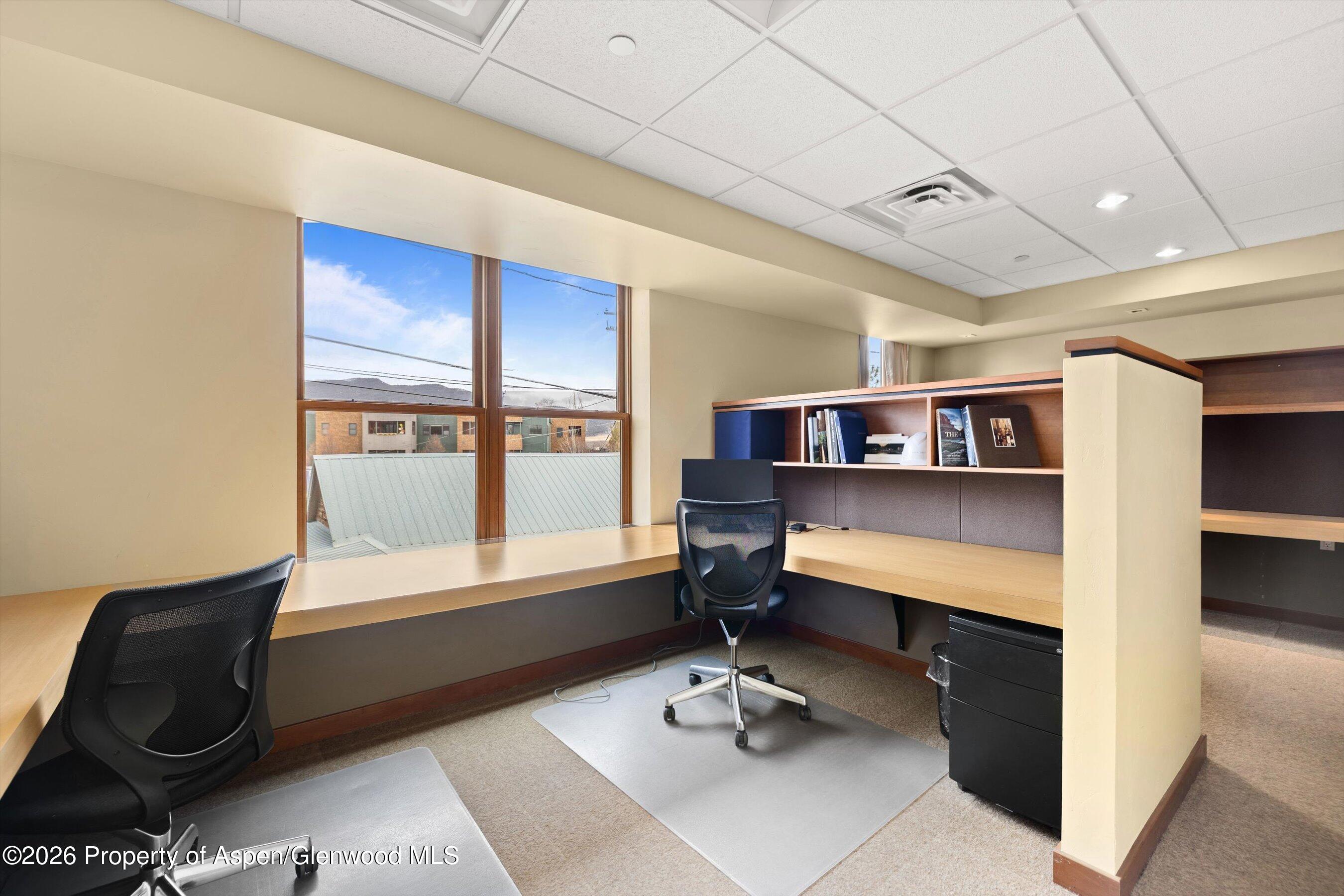 Office workspace with built-in desks, shelving, two black chairs, and large windows.