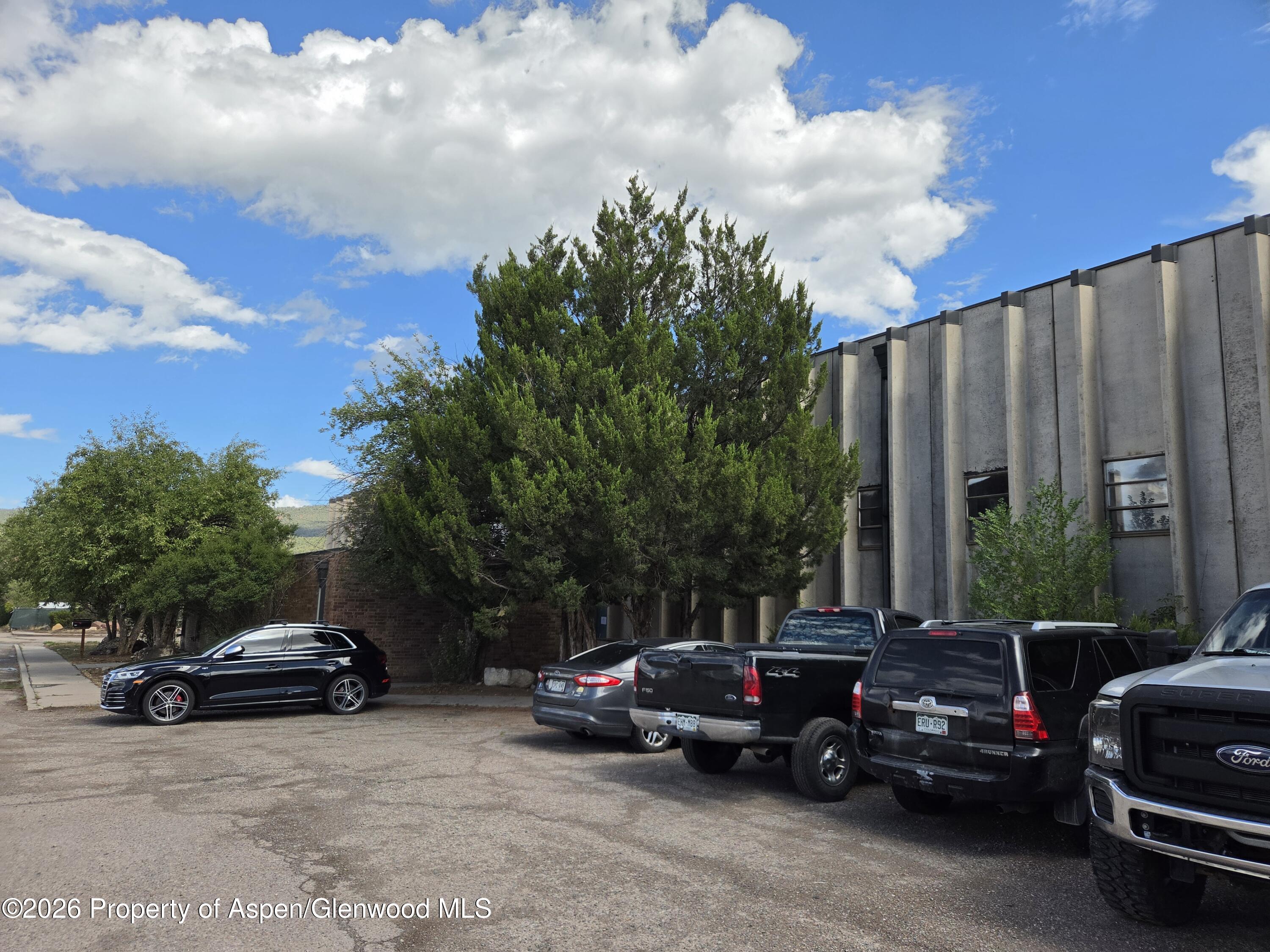 Gray commercial building beside a parking lot with parked vehicles and mature trees.
