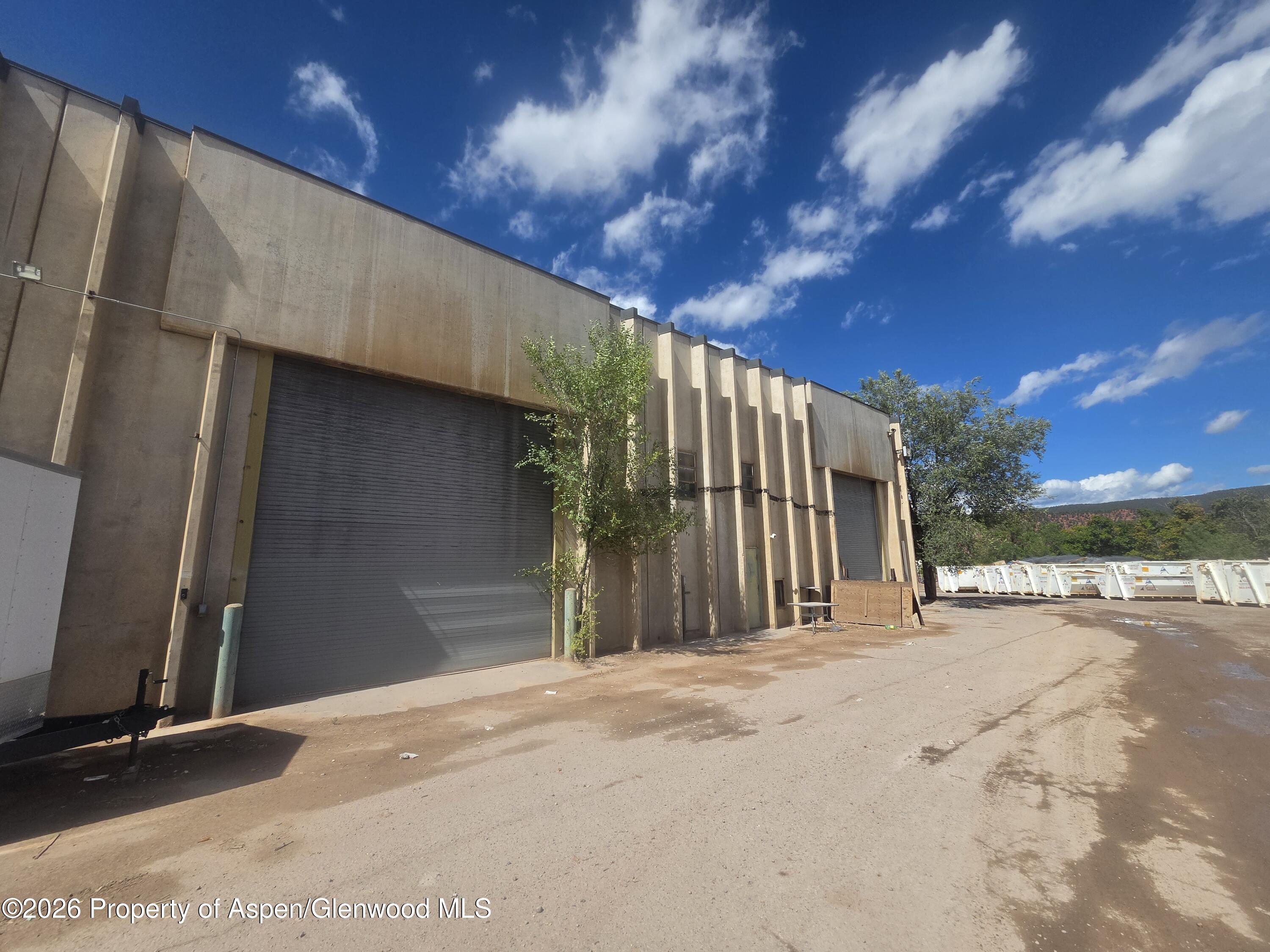 Industrial warehouse with two roll-up doors, a dirt service yard, and dumpsters beside trees and a mountain ridge.