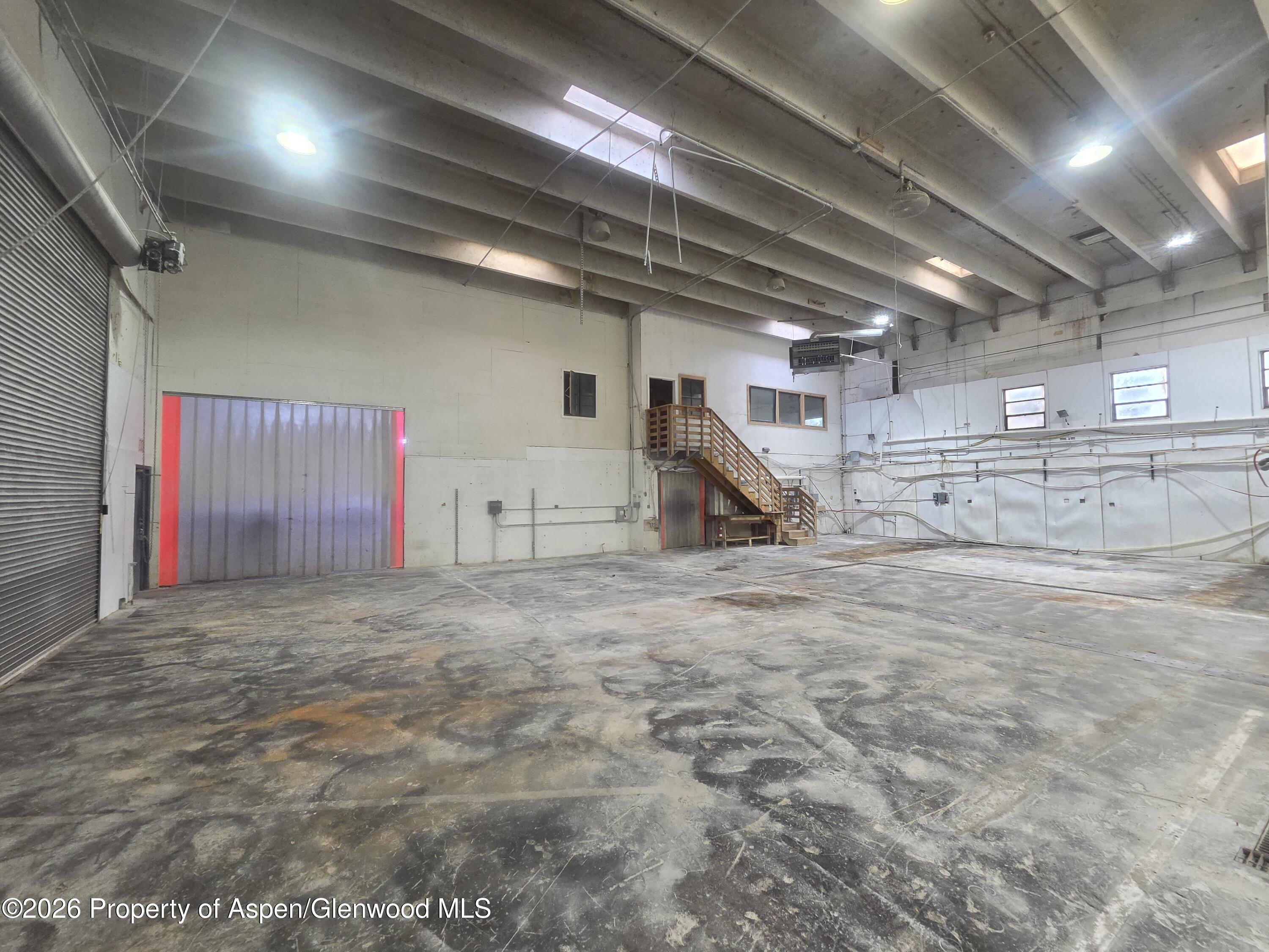 Vacant warehouse interior with a broad concrete floor, roll-up doors, and a wooden staircase.