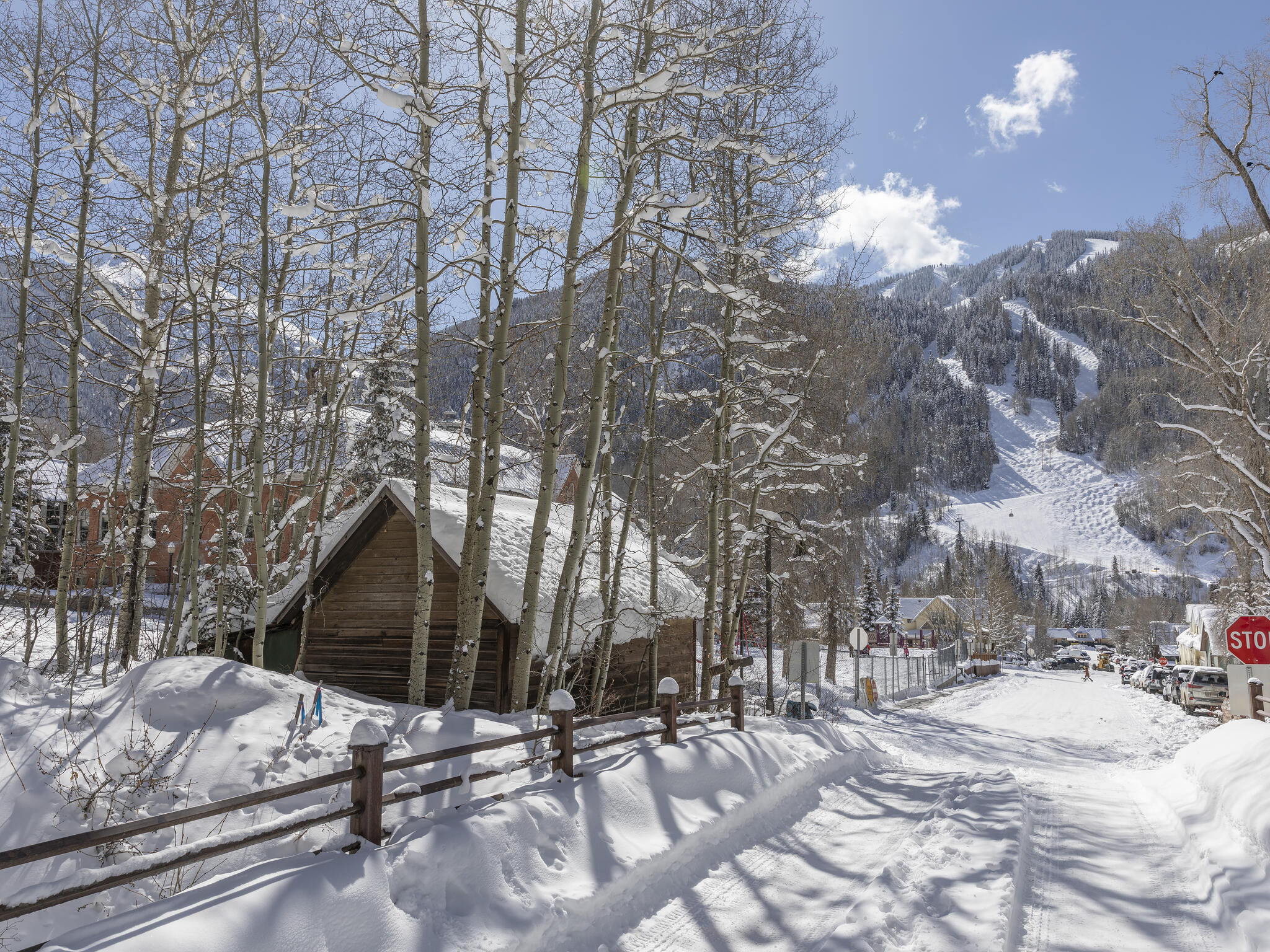 Snow-covered cabin among aspens beside a street, with a forested ski slope rising behind it.
