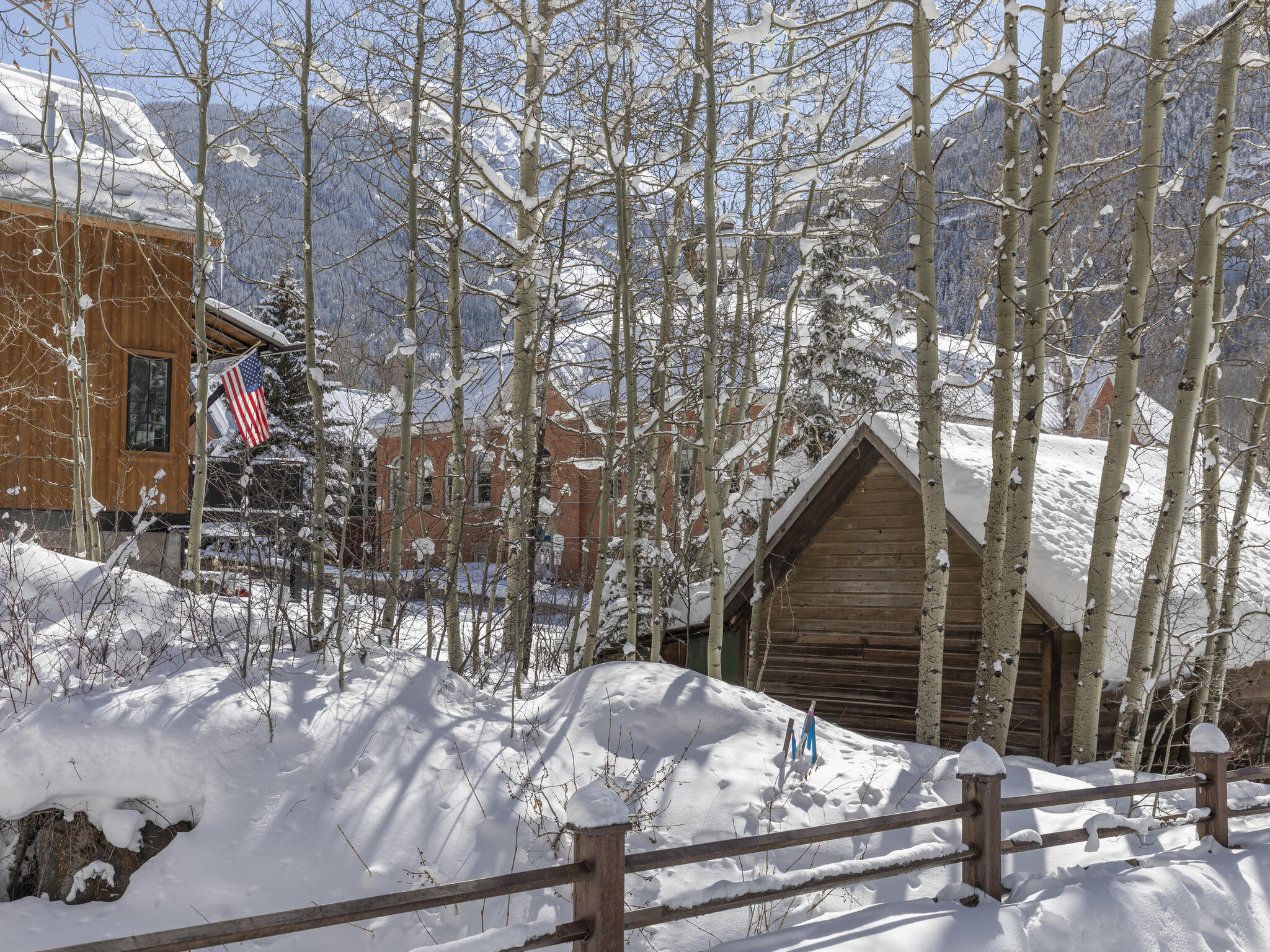 Wood-sided mountain homes among snow-covered aspens, with a wooden fence and mountain slopes beyond.