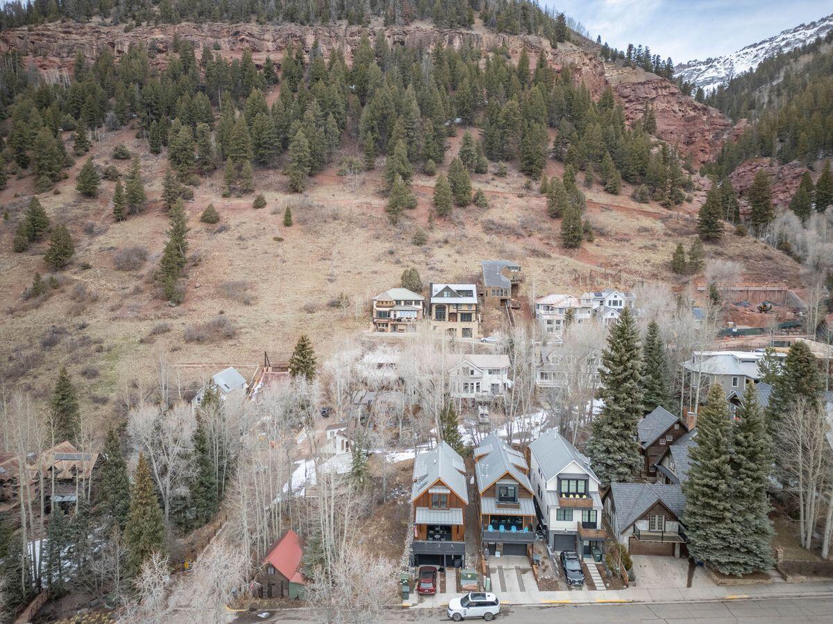 Aerial view of homes below a wooded hillside, with snow-covered mountains beyond.