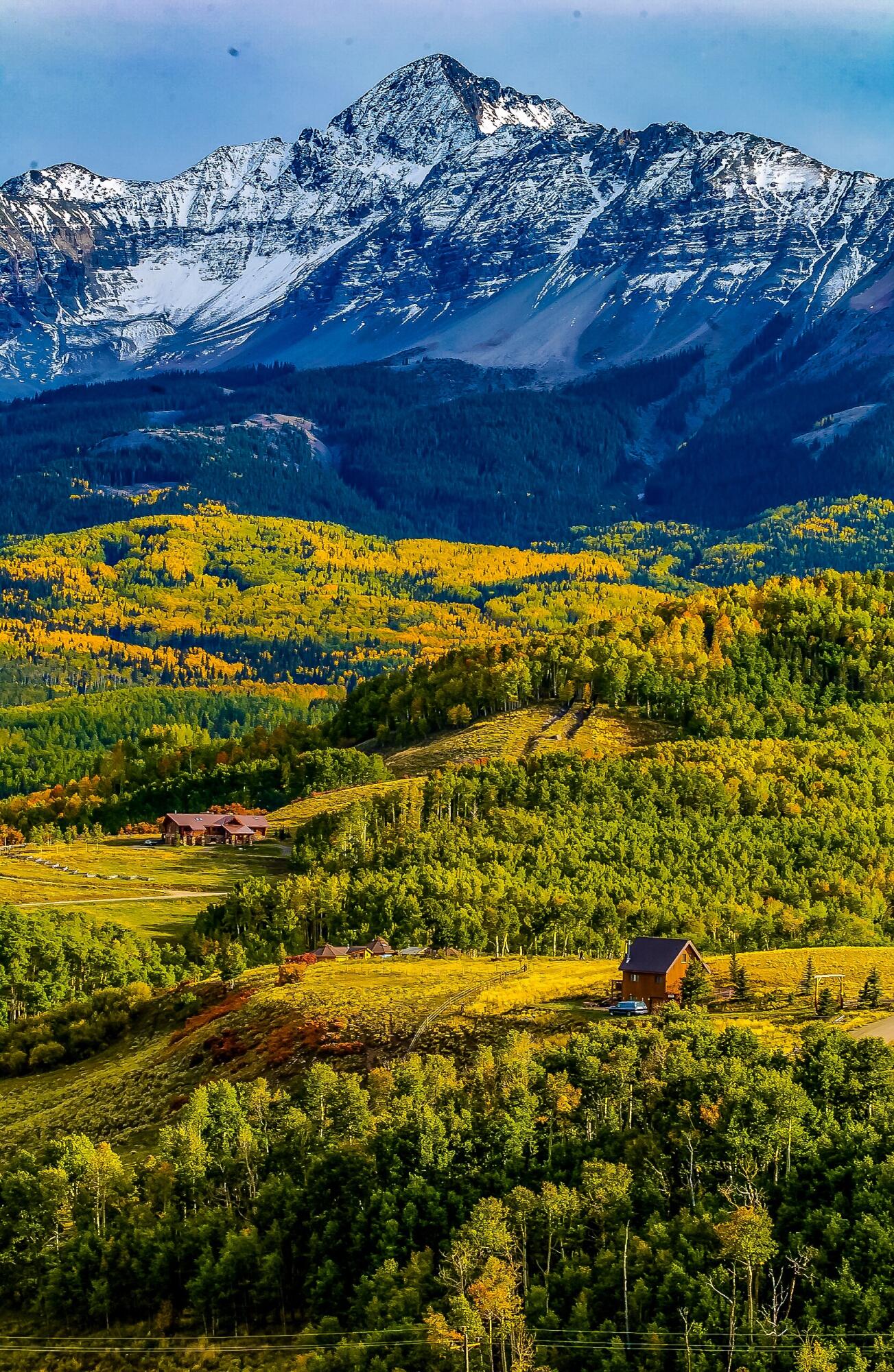 Snow-covered mountain above autumn-colored forest, rolling hills, and scattered homes.