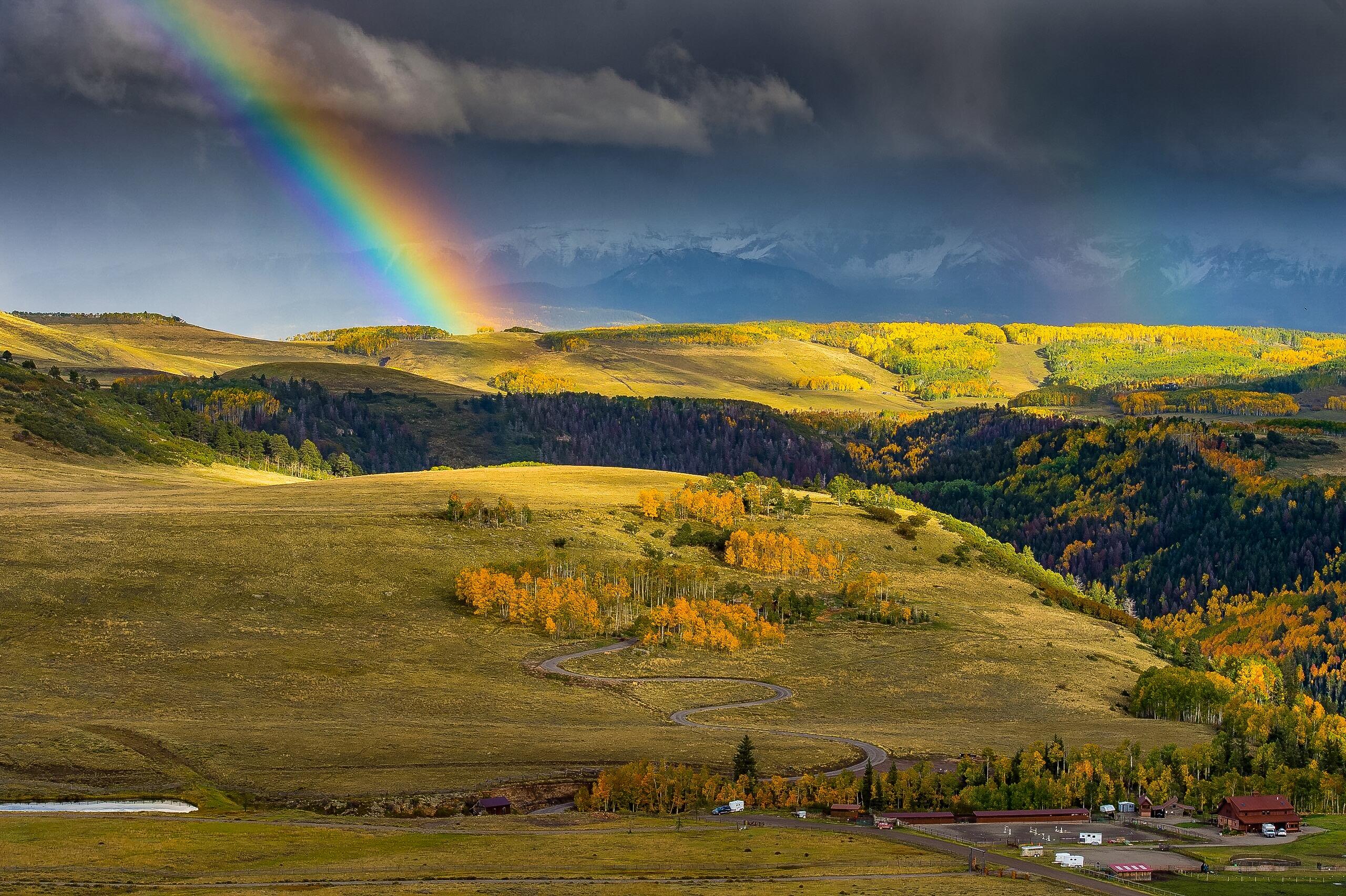 Rolling rural land with a winding road, golden autumn trees, distant mountains, and a rainbow.