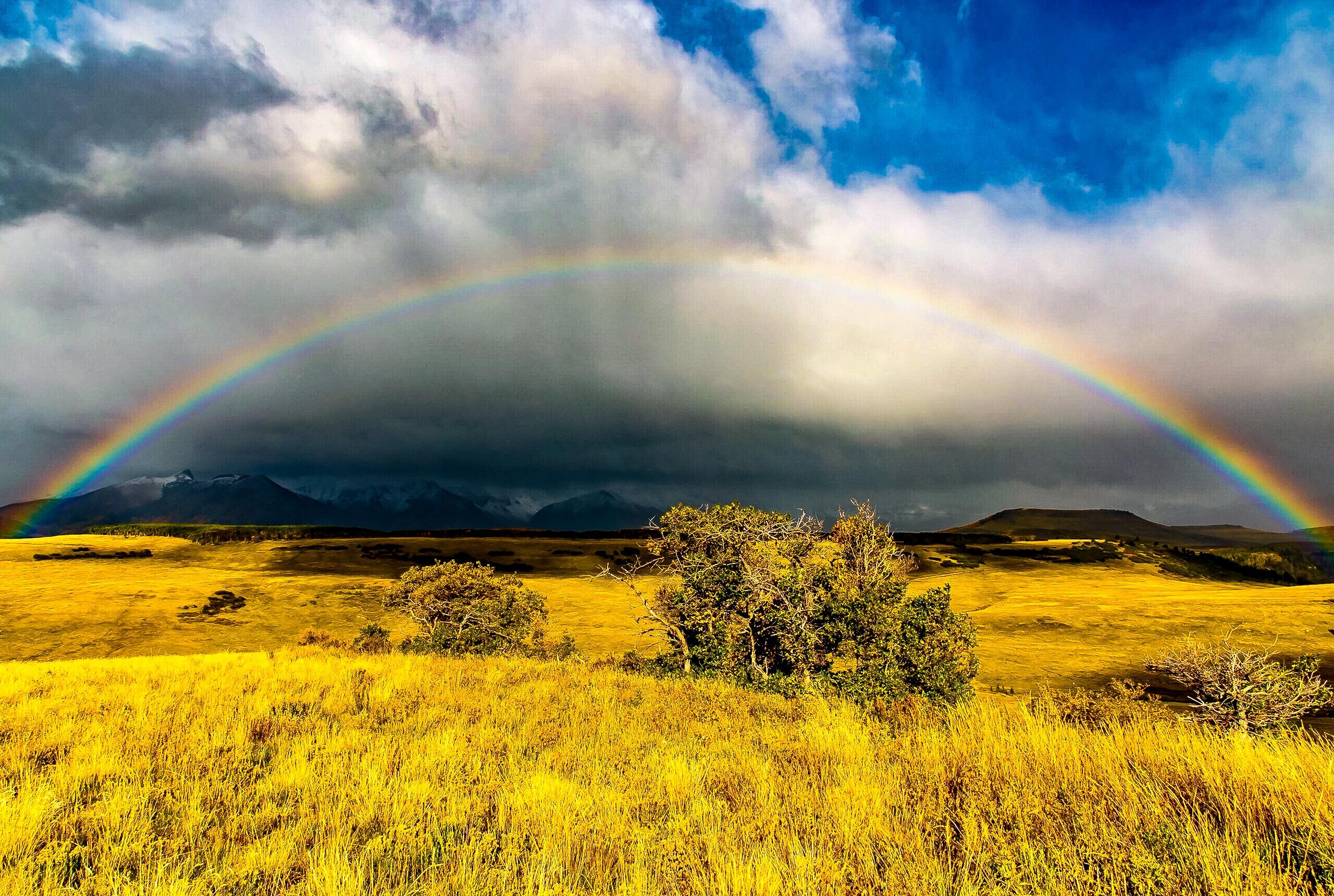 Golden grassland and mountain ridges beneath a dramatic rainbow and storm clouds.
