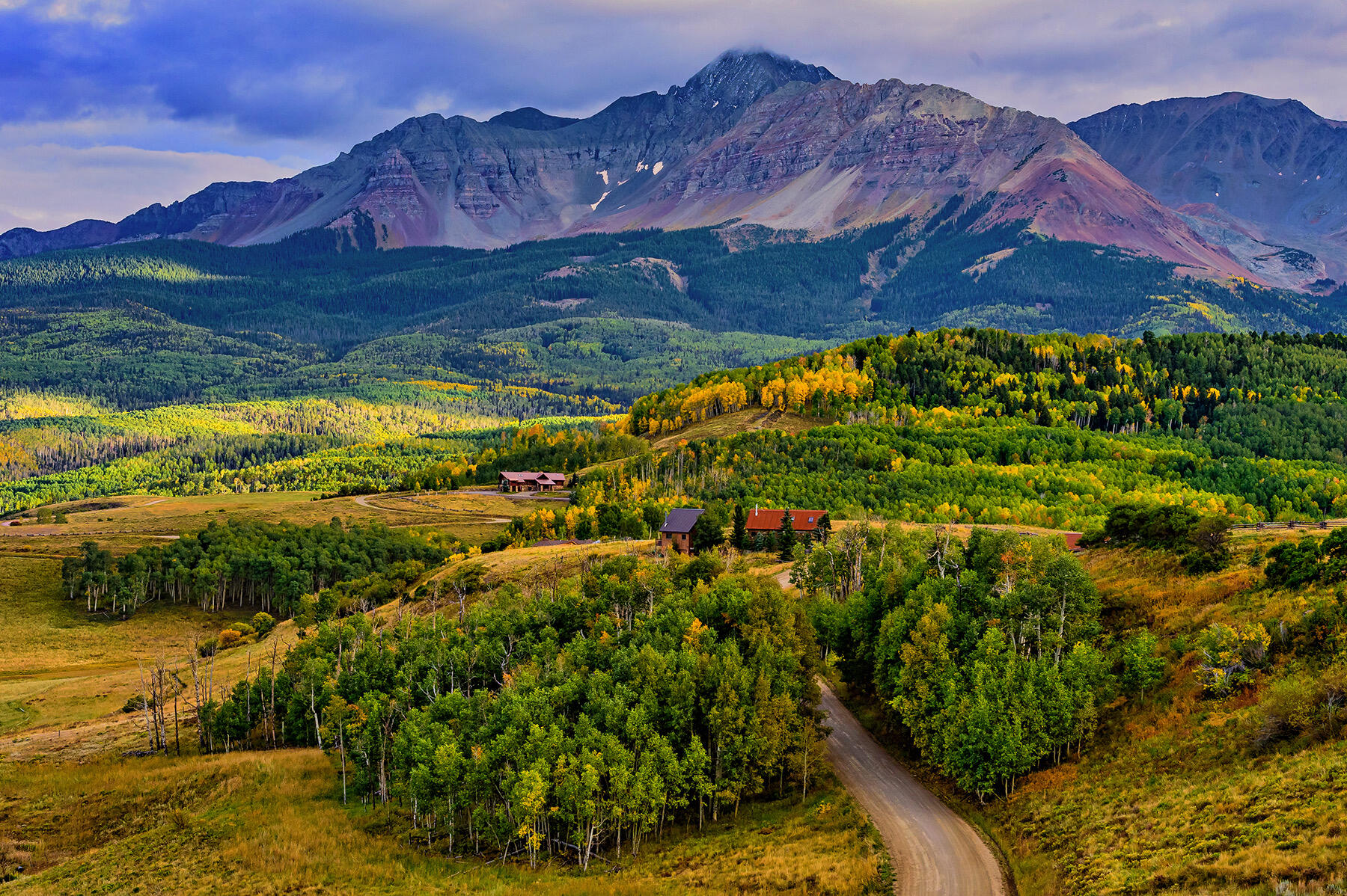 Mountain property with scattered buildings and a winding road amid autumn forest and meadows.