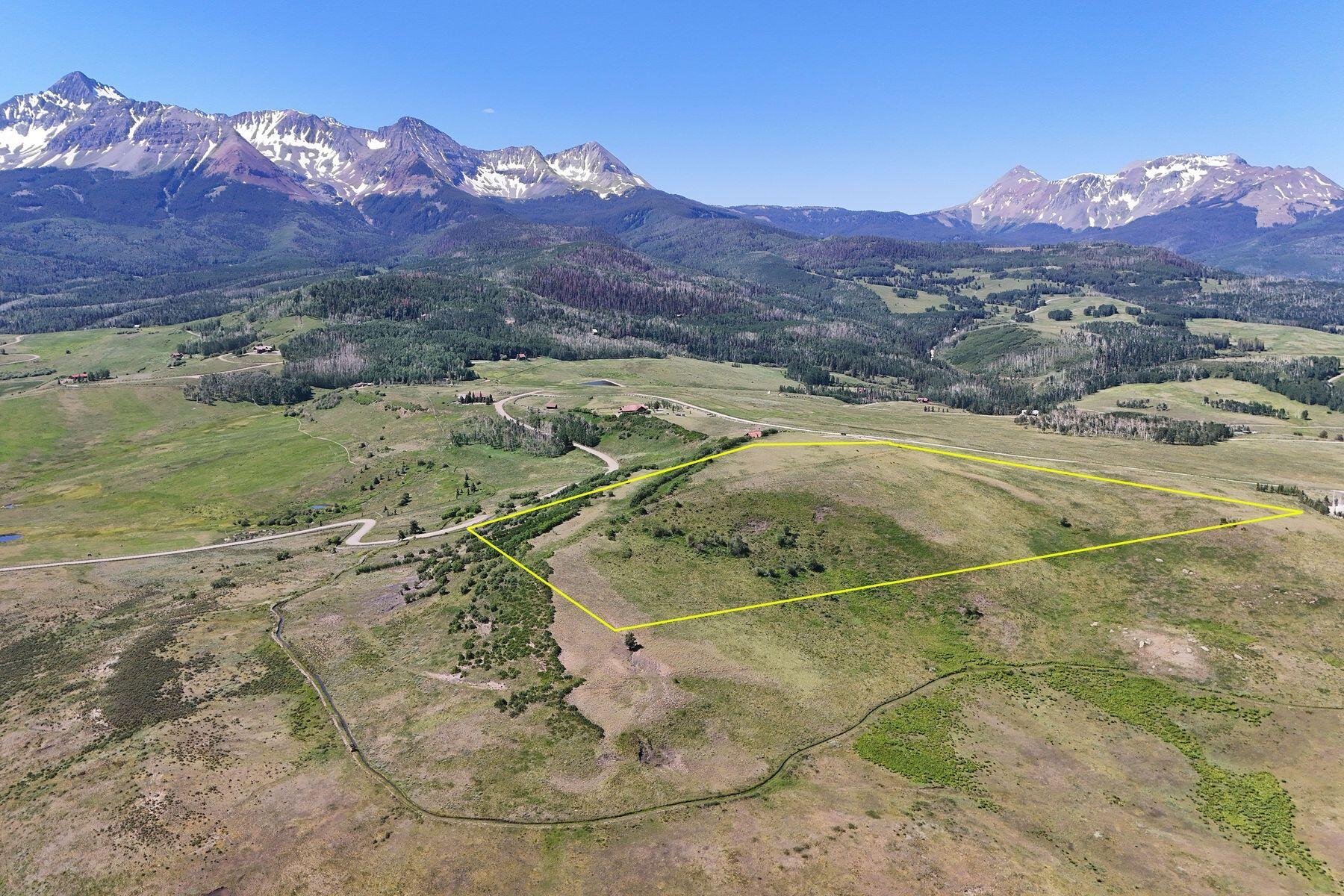 Aerial view of a rural parcel outlined in yellow, with open grassland and snow-streaked mountains beyond.