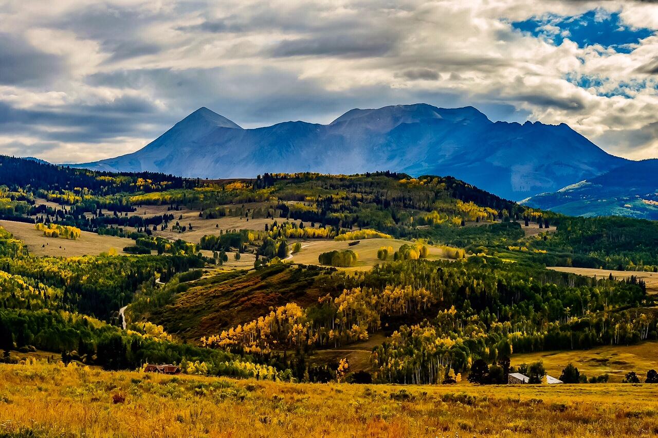 Rolling autumn fields and golden trees stretch toward a broad mountain range beneath dramatic clouds.