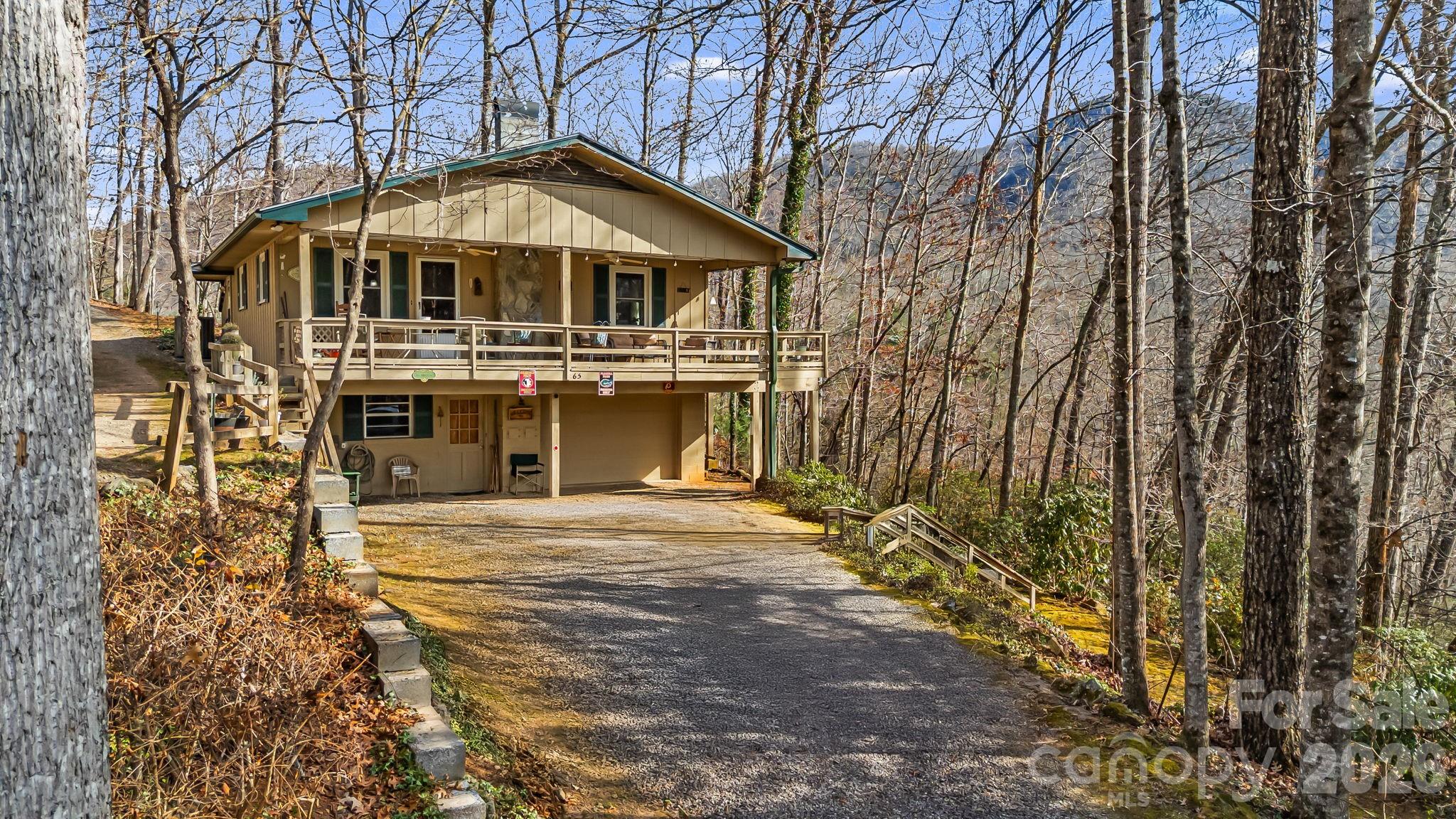 Raised tan home with a broad wooden deck, gravel driveway, and wooded surroundings.