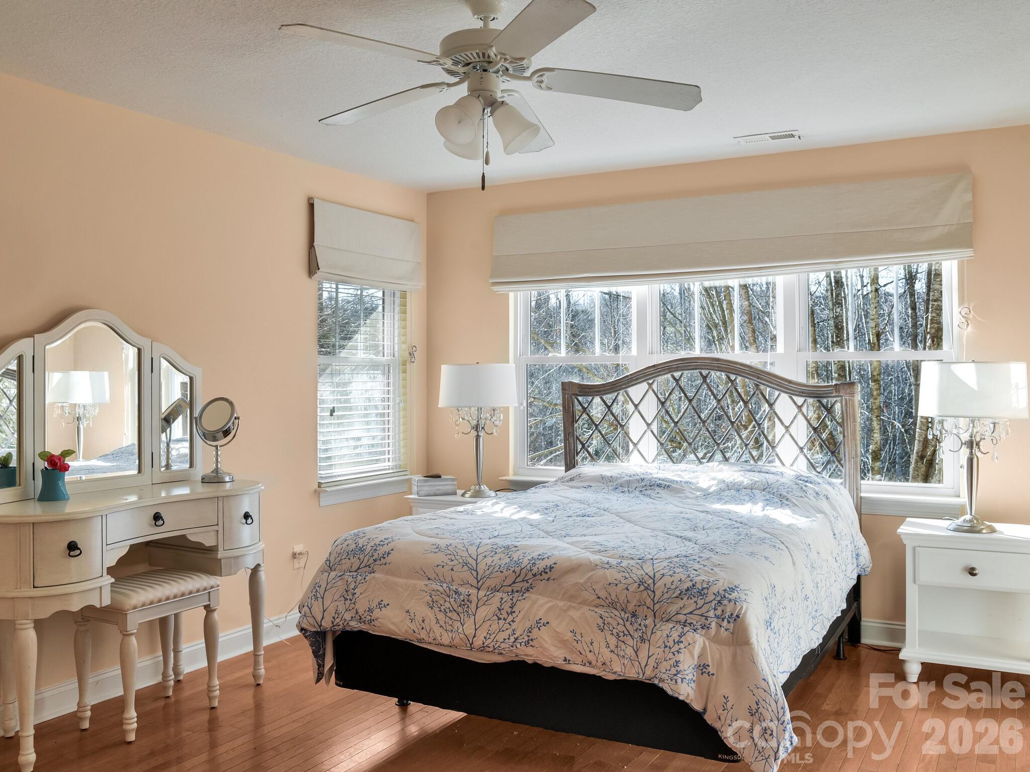 Bedroom with a patterned quilt, white vanity, and broad windows overlooking bare trees.