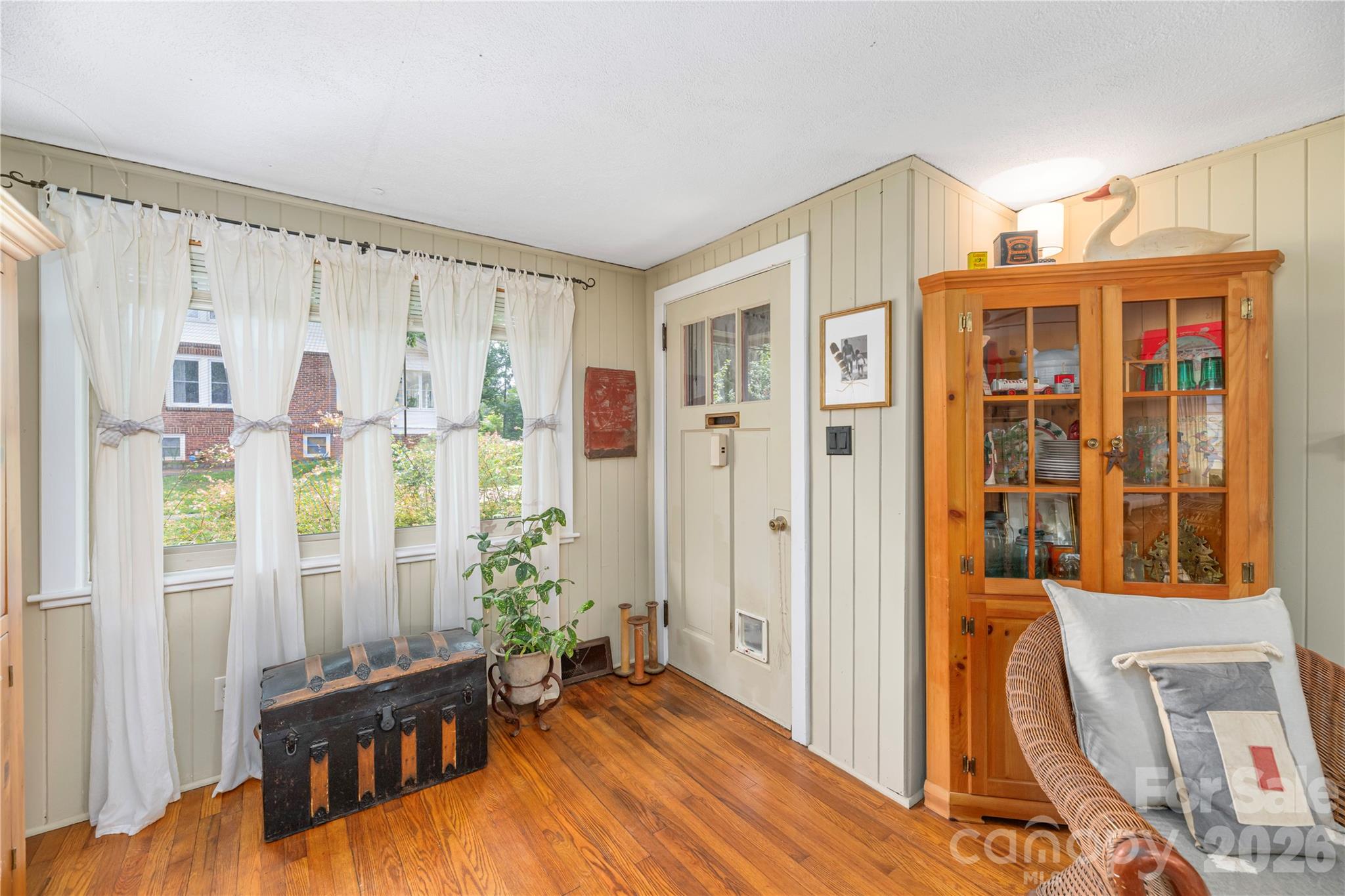 Furnished room with hardwood floors, a curtained window, antique trunk, and wooden display cabinet.