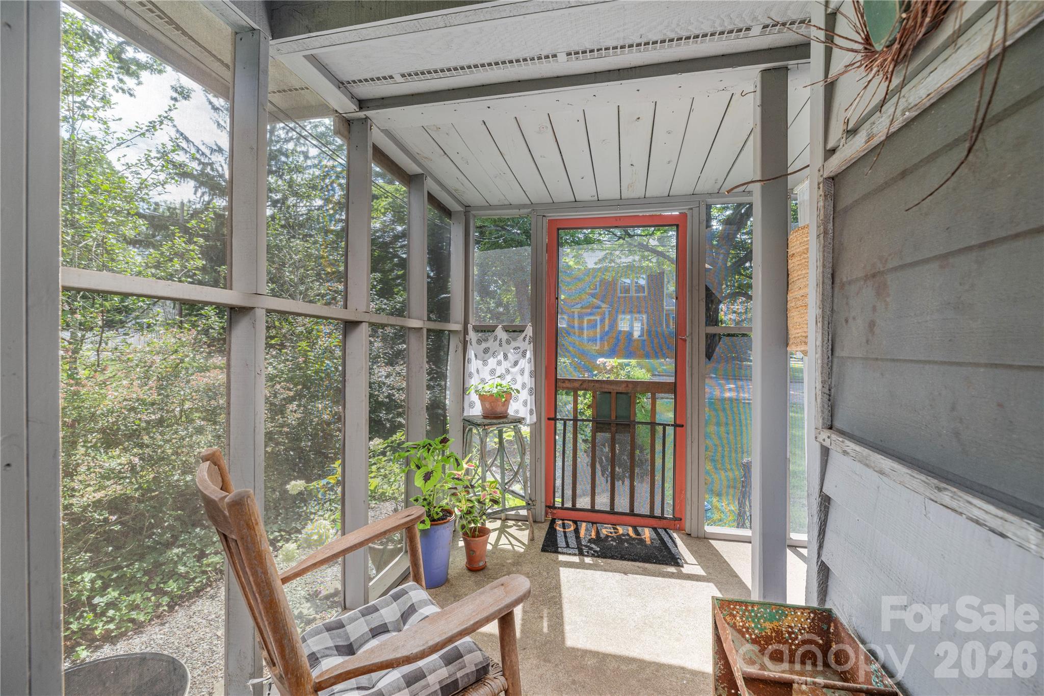 Enclosed porch with a red-framed screen door, wooden chair, potted plants, and views of trees.