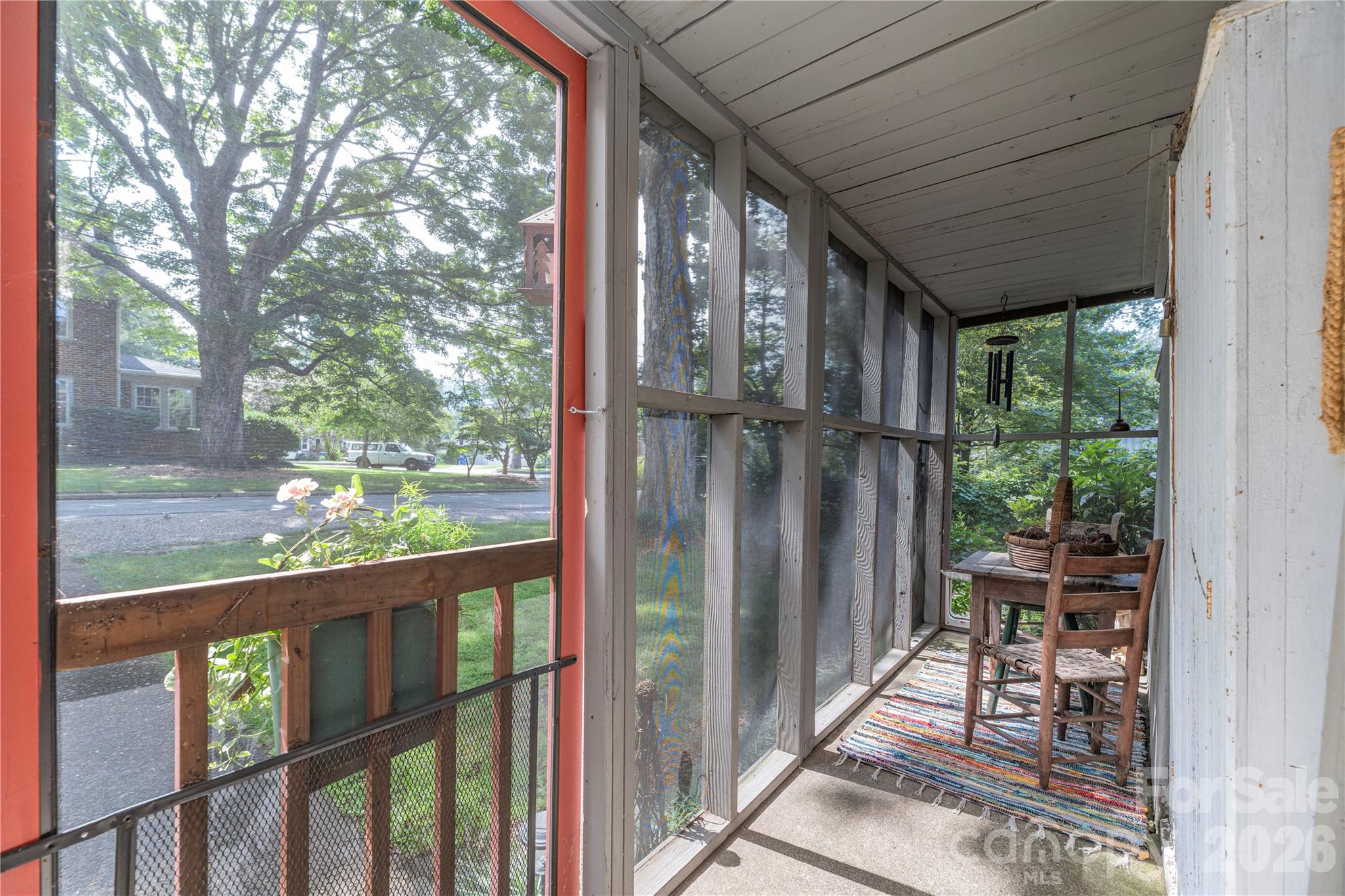Screened porch with wooden chairs, a striped rug, and leafy street views.