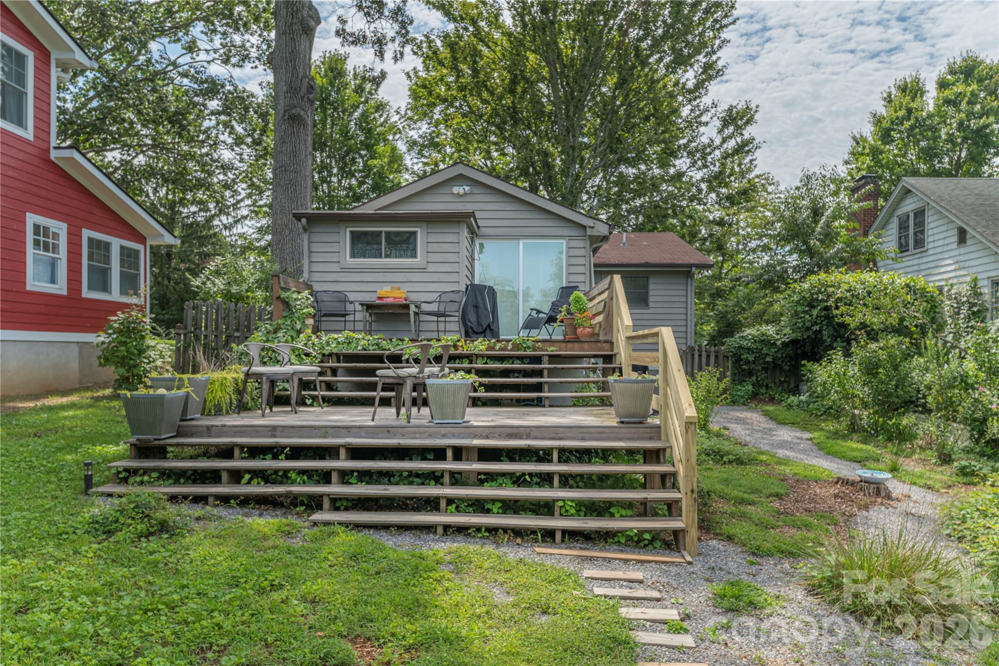 Gray house with a wide, tiered wooden deck furnished with patio chairs and surrounded by a leafy yard.