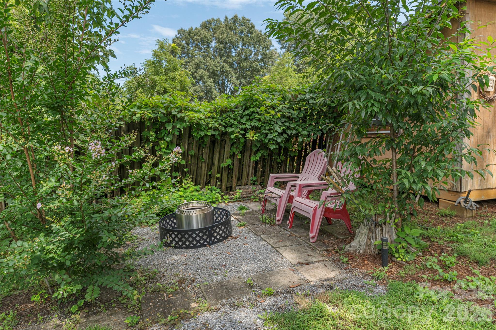 Backyard patio with a metal fire pit, pink chairs, gravel, and dense greenery.