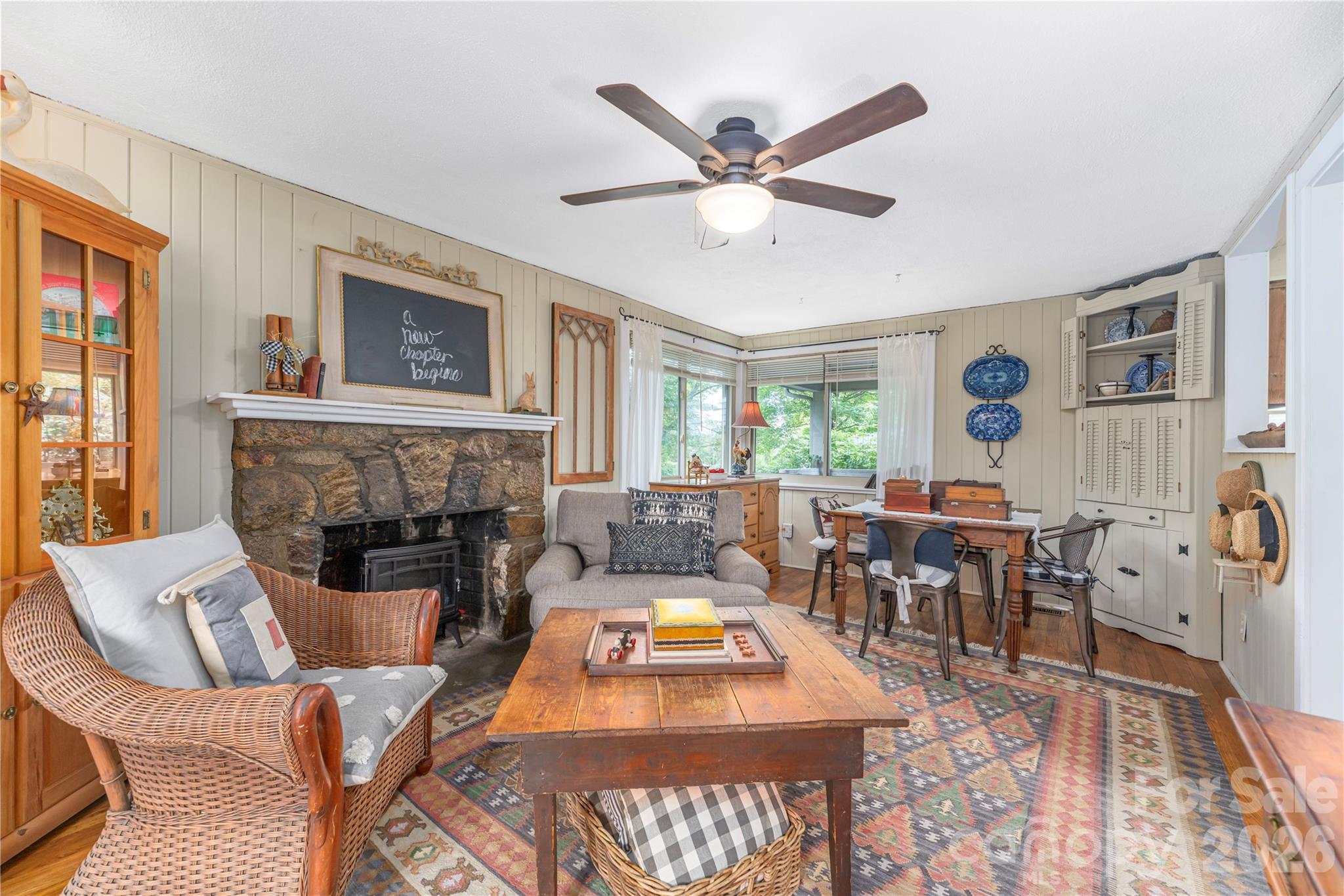 Living and dining room with a stone fireplace, wicker seating, and a bright bay window.