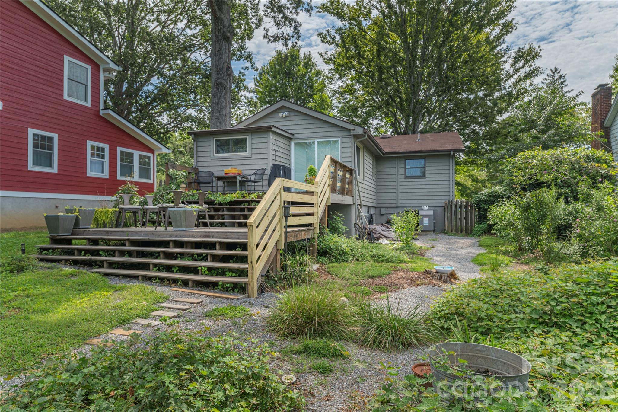 Gray house with a raised wooden deck and stairs overlooking a leafy backyard and gravel path.