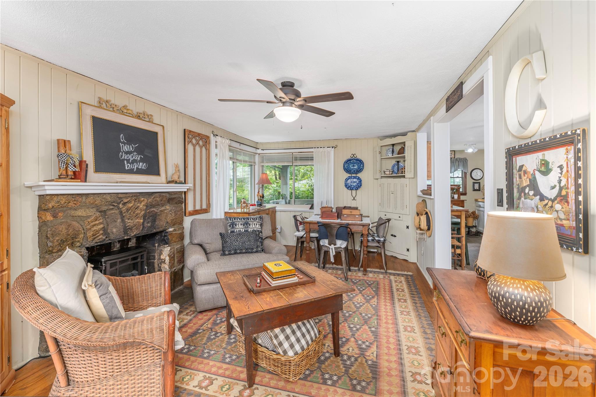 Living room with a stone fireplace, wicker chair, and dining area beside a bay window.