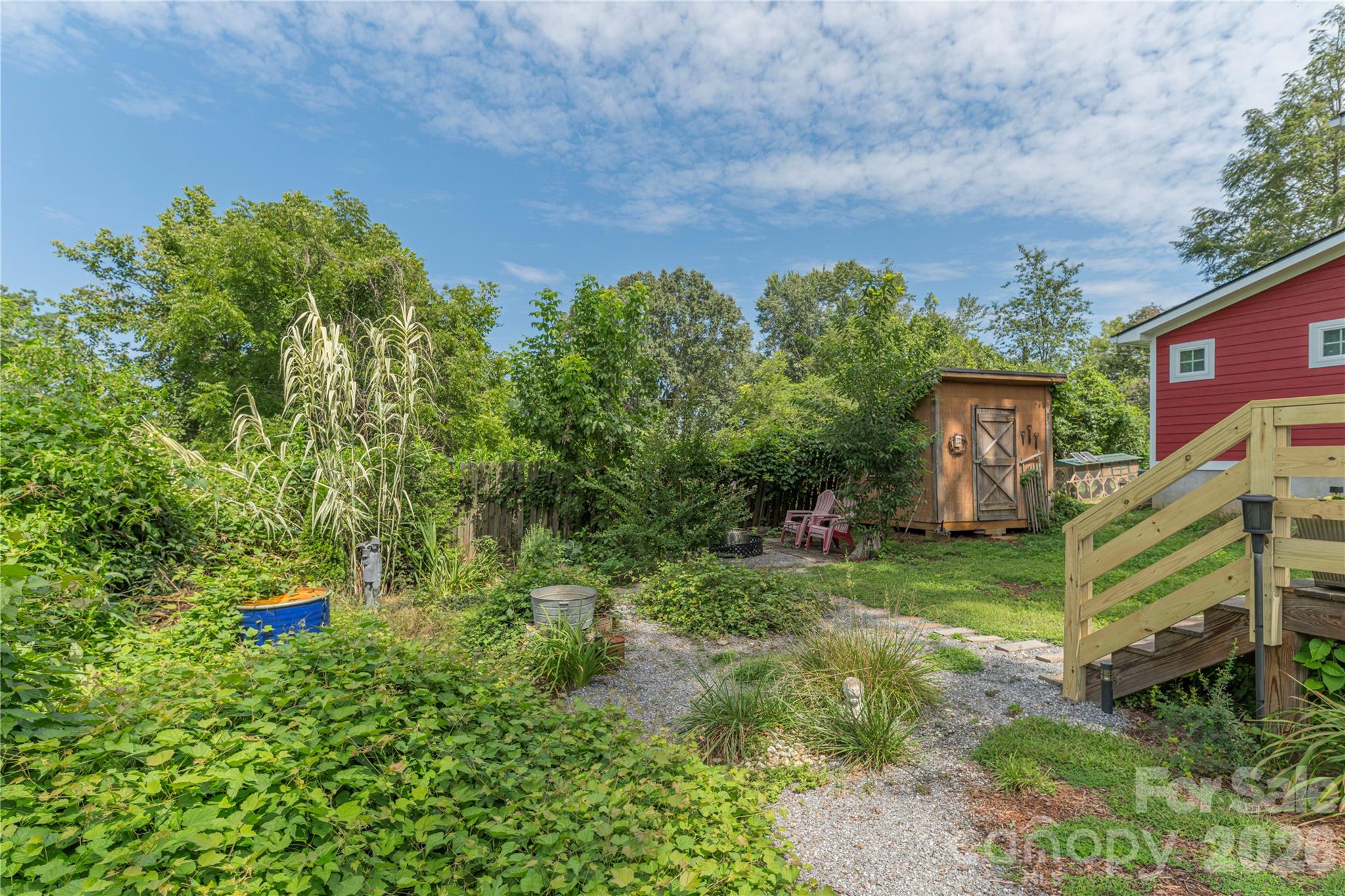 Backyard with a gravel path, wooden shed, dense garden, and stairs beside a red house.
