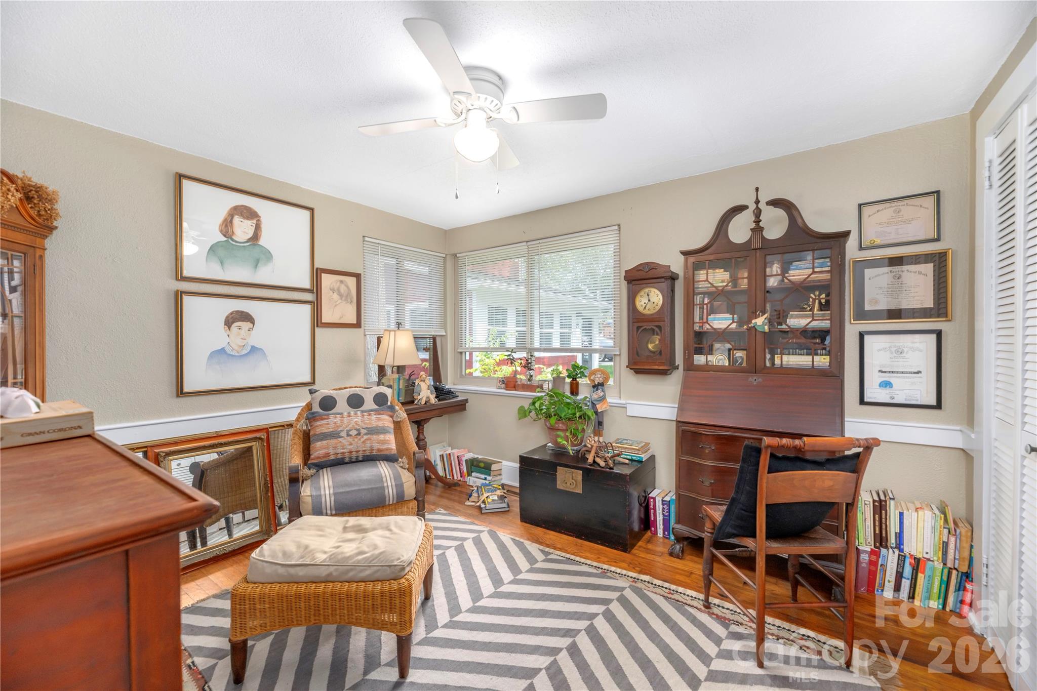 Furnished home office with a wooden secretary desk, glass-front bookcase, corner windows, and patterned rug.