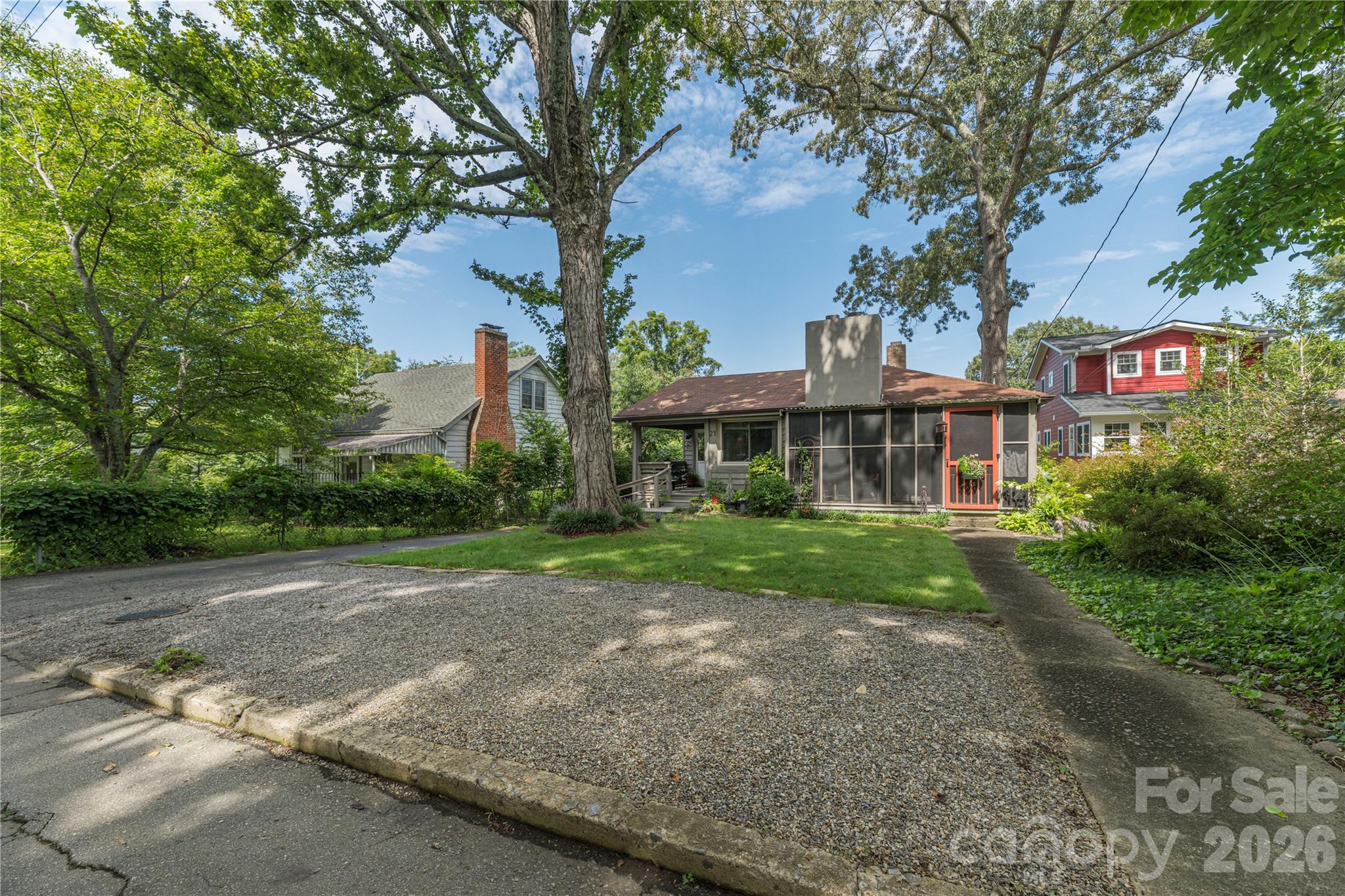Single-story house with a screened porch and red door, shaded by mature trees beside a gravel parking area.