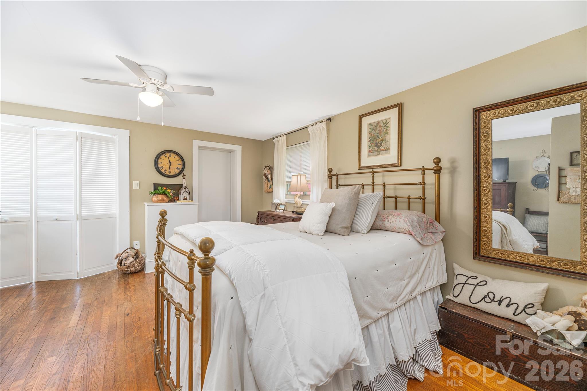 Furnished bedroom with a brass bed, white bedding, wood floors, and an ornate wall mirror.