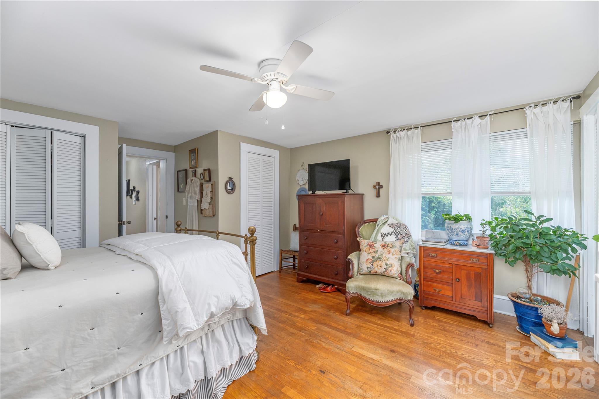 Furnished bedroom with a white bed, warm hardwood floors, large curtained windows, and a wooden dresser.