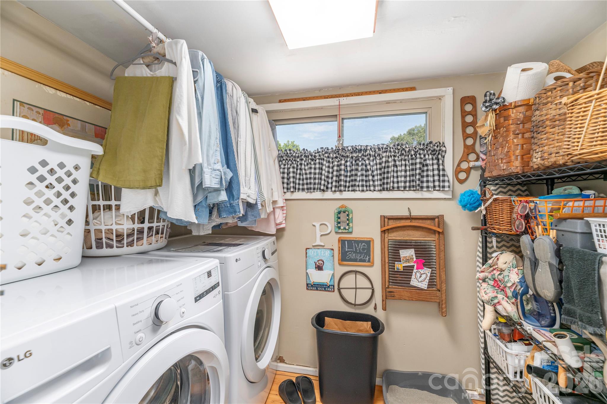 Laundry room with washer and dryer, hanging clothes, a curtained window, and shelves of baskets.