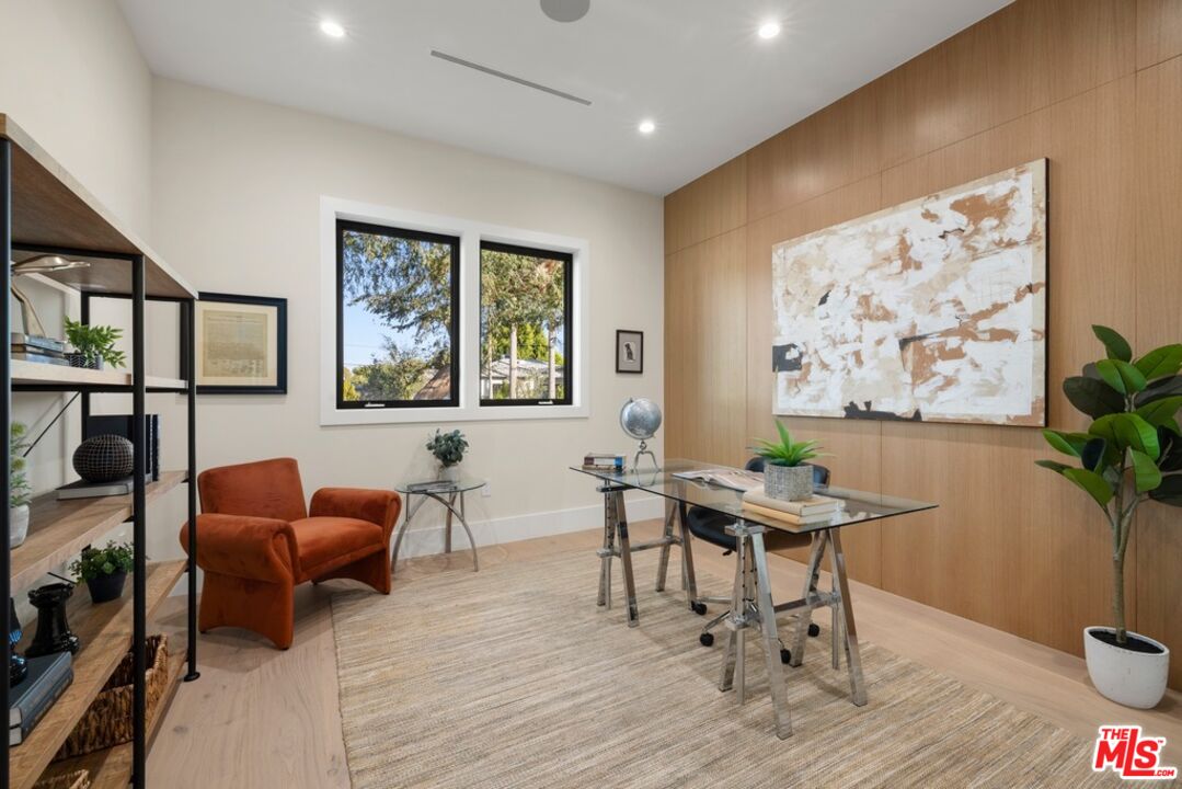 Furnished home office with a glass desk, orange armchair, wood-paneled wall and tree views through large windows.