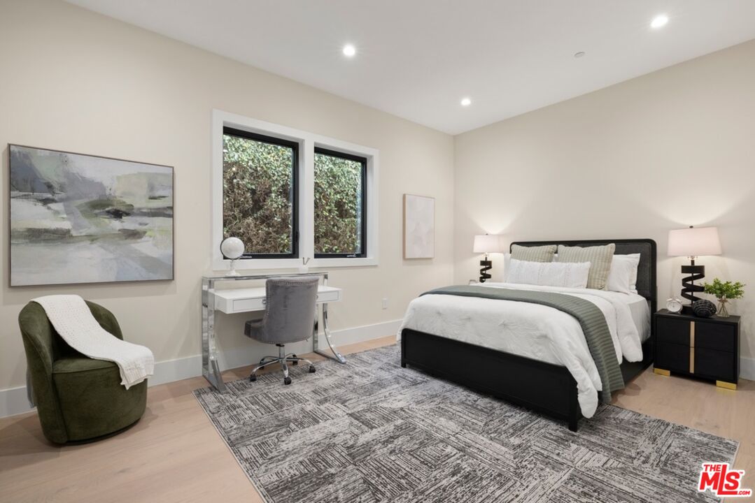 Staged bedroom with a black bed, white bedding, desk beneath two windows, and patterned rug.