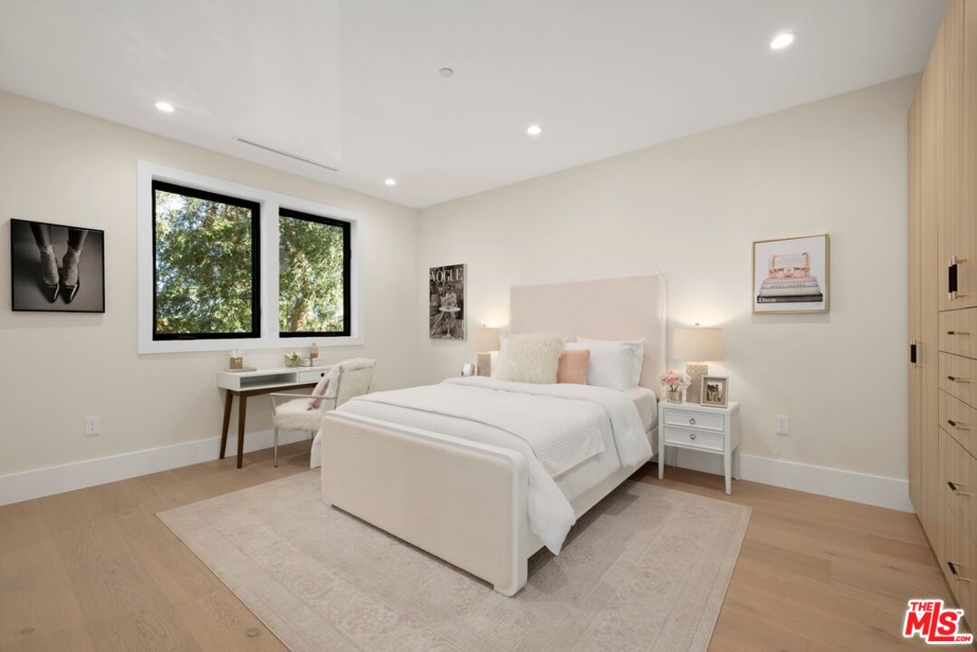 Staged bedroom with a white upholstered bed, leafy views through two windows, and built-in wood cabinetry.