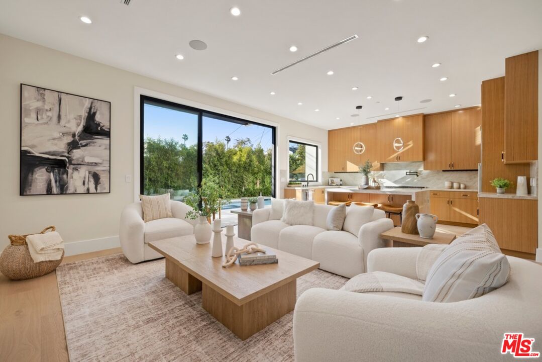 Open-plan living room with white sofas, warm wood kitchen cabinetry, and sliding doors to greenery.